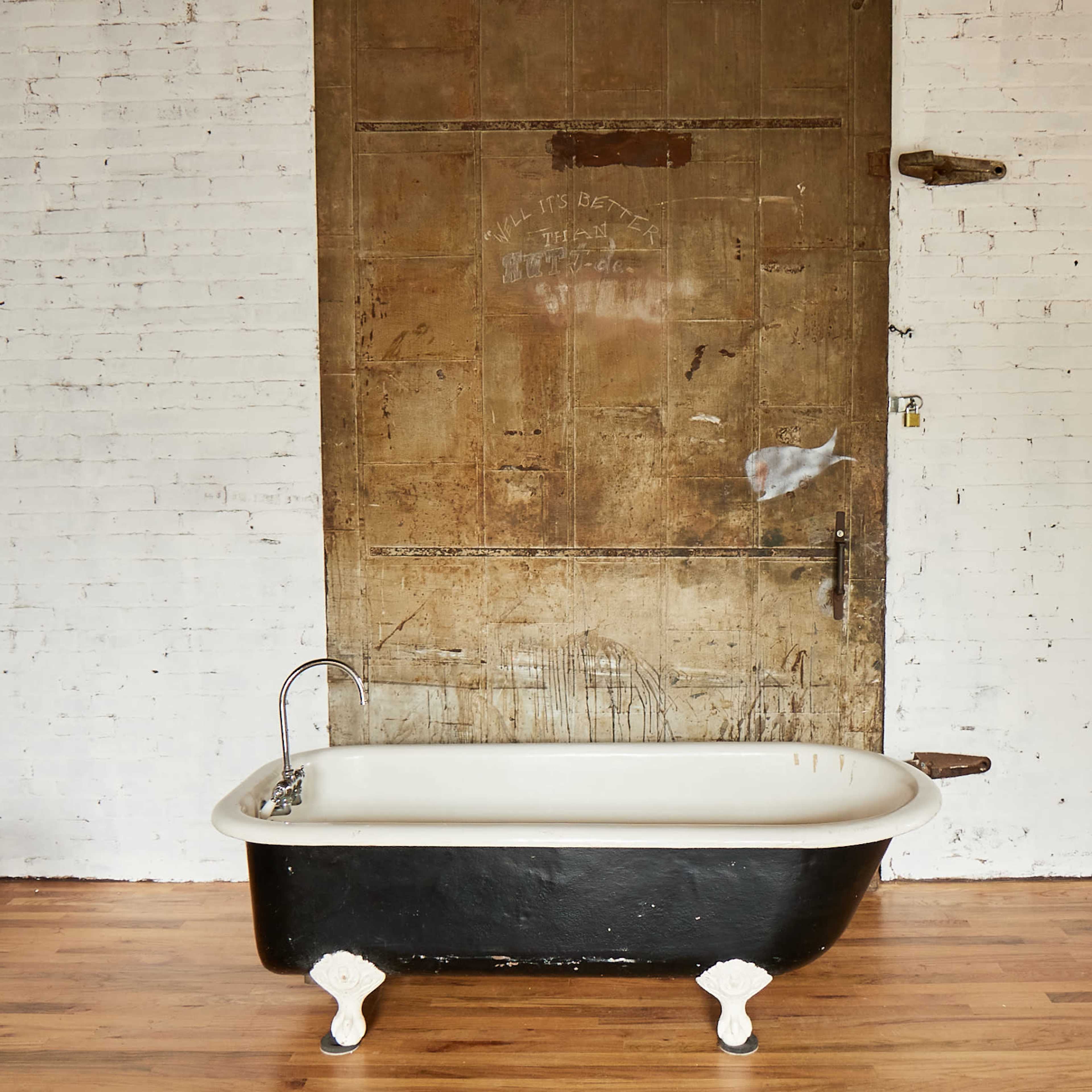 A black and white freestanding bathtub is positioned in front of a large wooden door against a white brick wall.