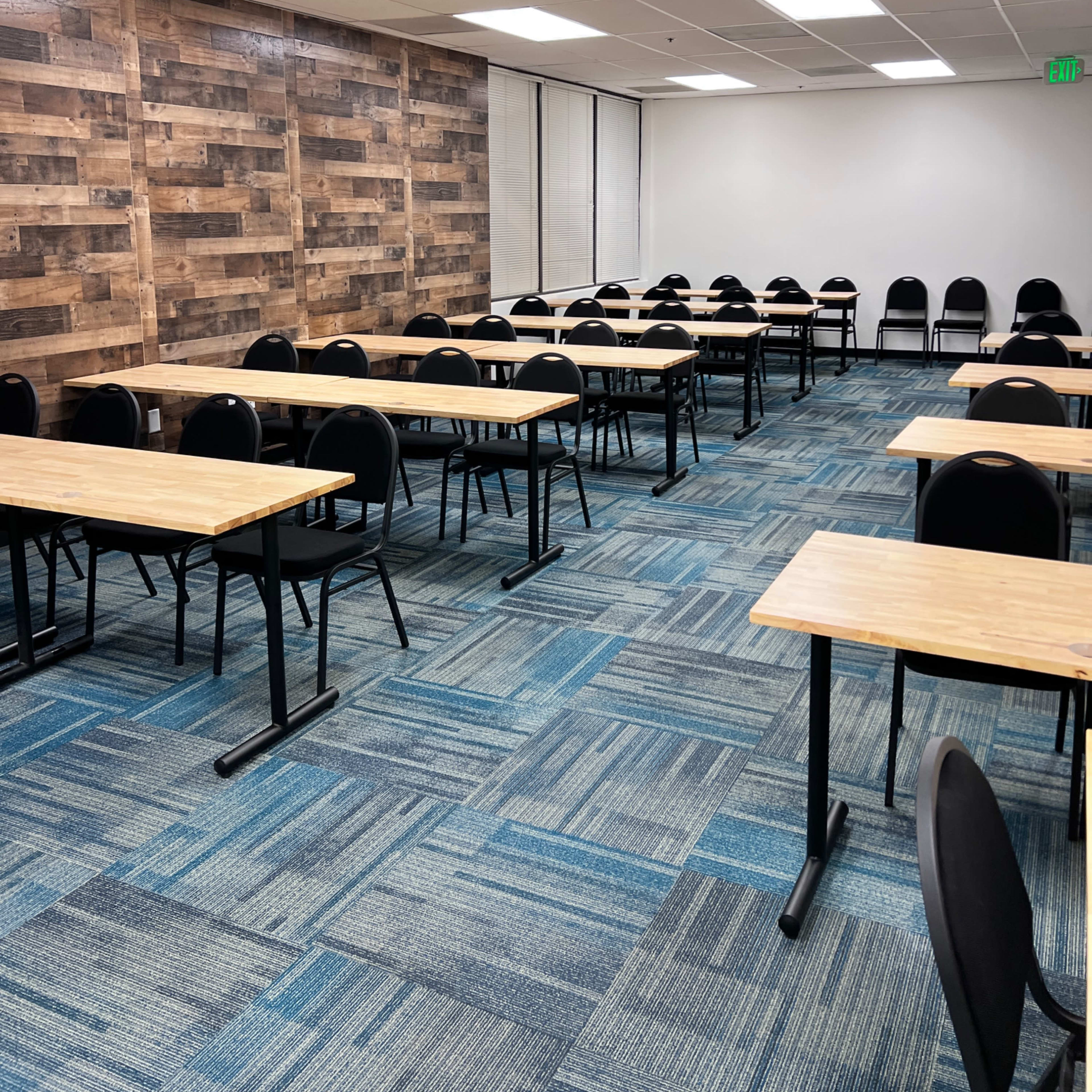 A spacious classroom arranged with rows of wooden tables and black chairs on a blue carpet.