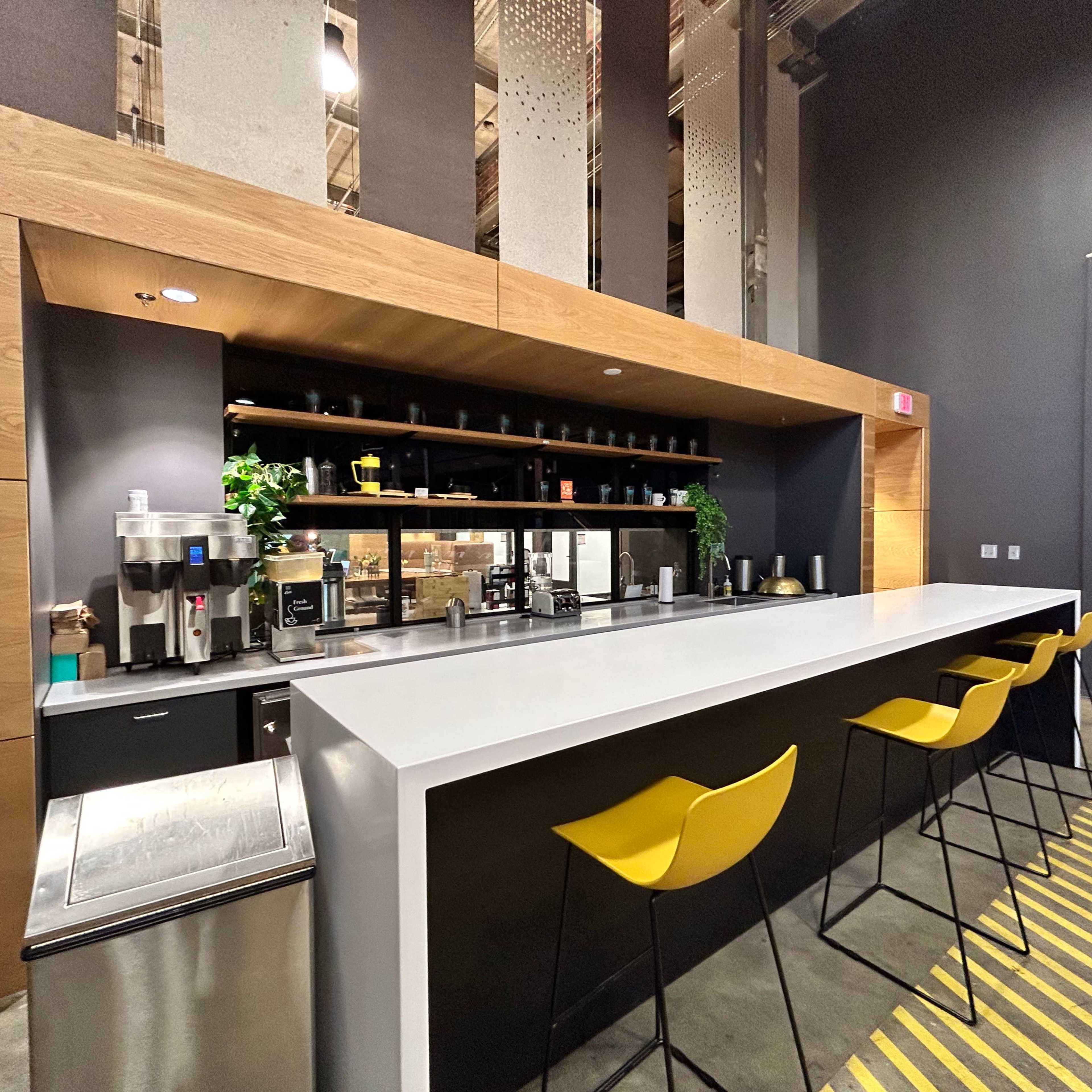 A modern kitchen area with a long, white countertop, yellow bar stools, and shelves displaying various kitchen items against a dark-colored wall.