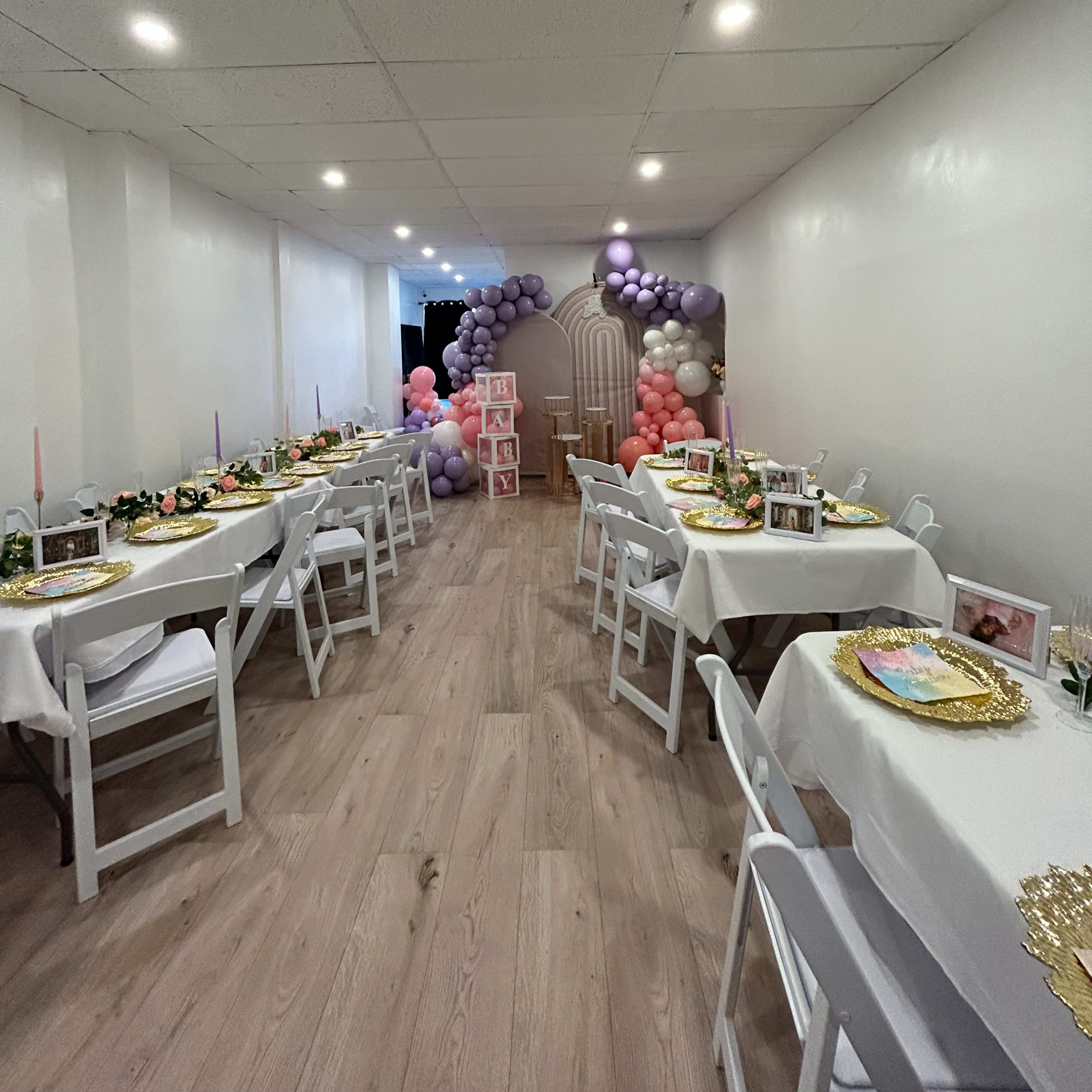 A spacious event room decorated with tables set for a celebration, featuring floral centerpieces and a balloon arch in the background.
