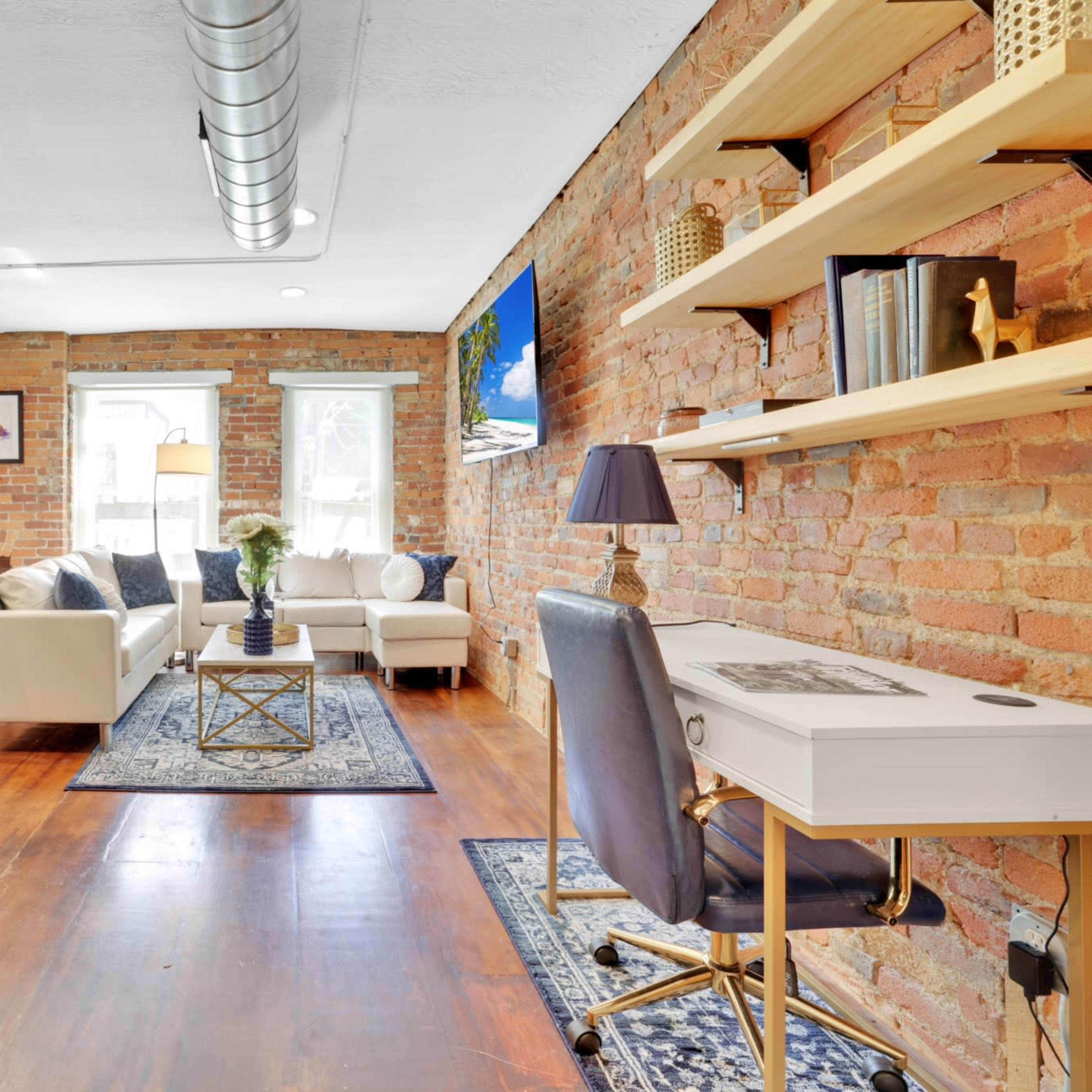 A bright living area with exposed brick walls, a sofa, a desk, and a decorative shelf, all arranged on hardwood flooring.