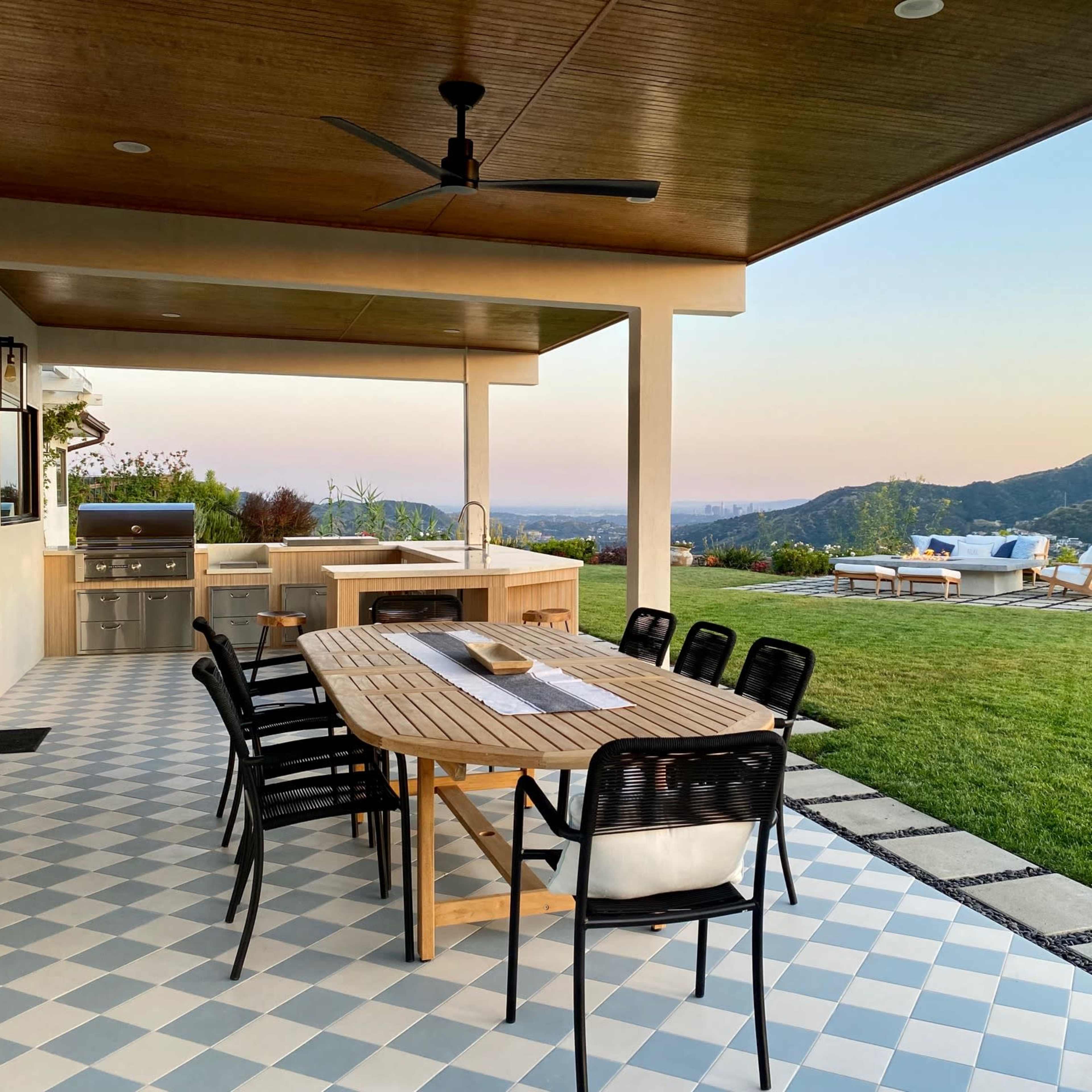 A patio features a large wooden dining table surrounded by black chairs, overlooking a lush green landscape and mountains under a clear sky.
