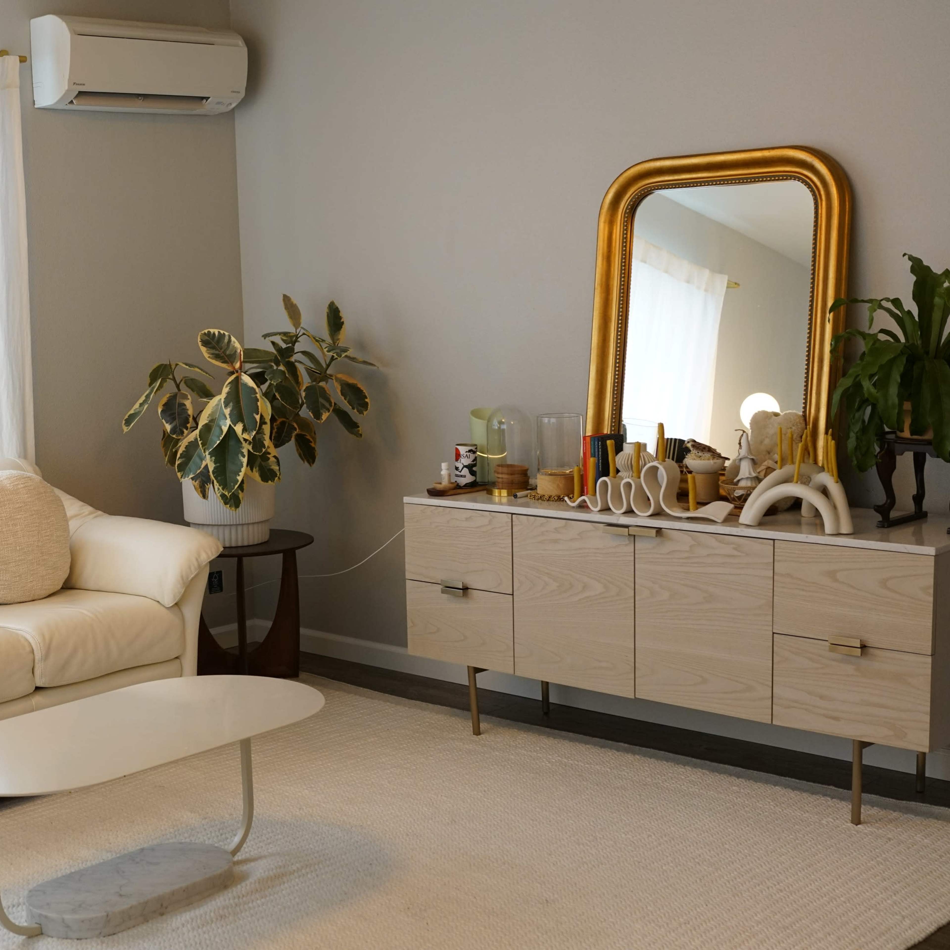 A living room with a light-colored sofa, a white coffee table, and a wooden sideboard, accompanied by a large mirror and a potted plant.