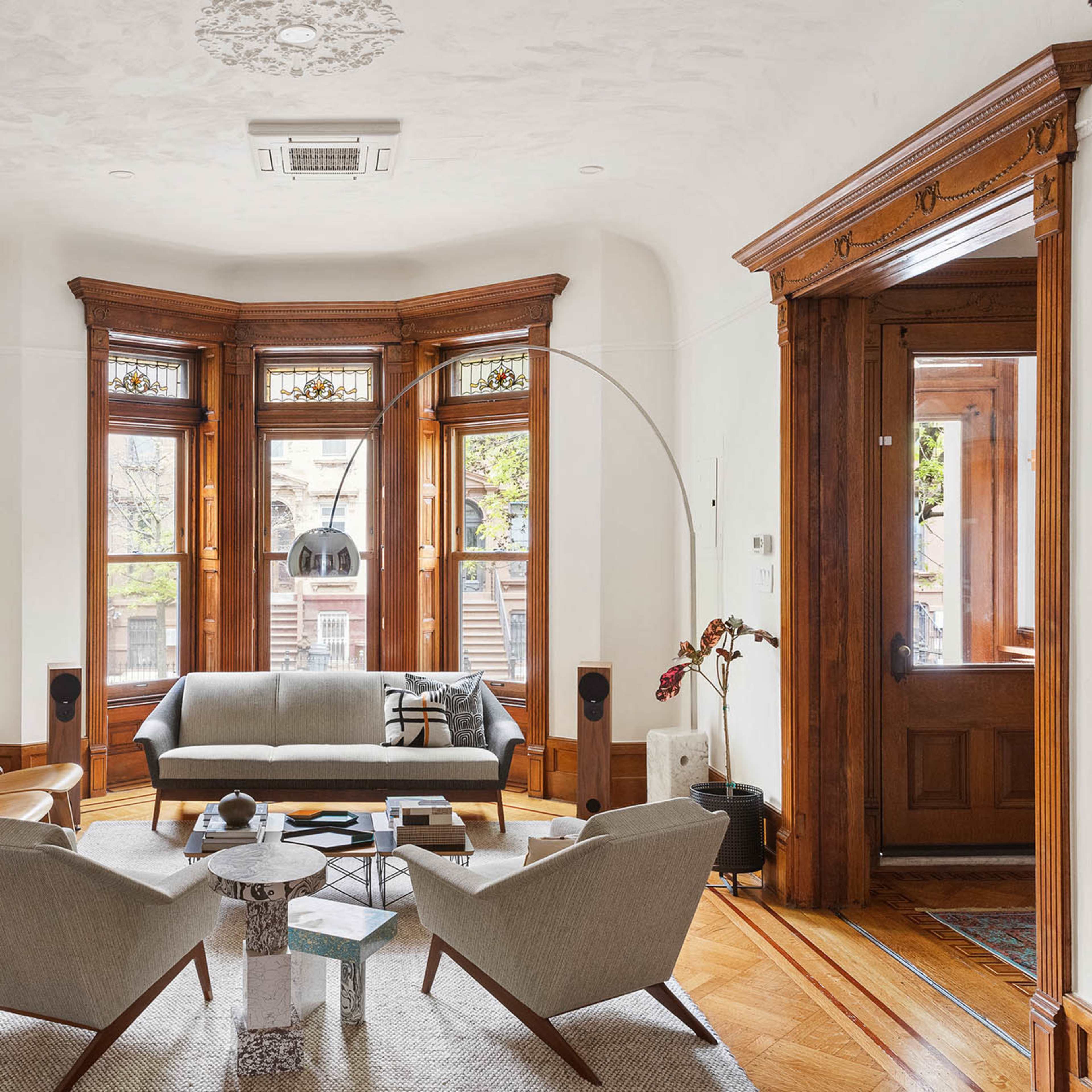 A living room with large bay windows, brown wooden trim, a gray sofa, two armchairs, and a record player on a table.