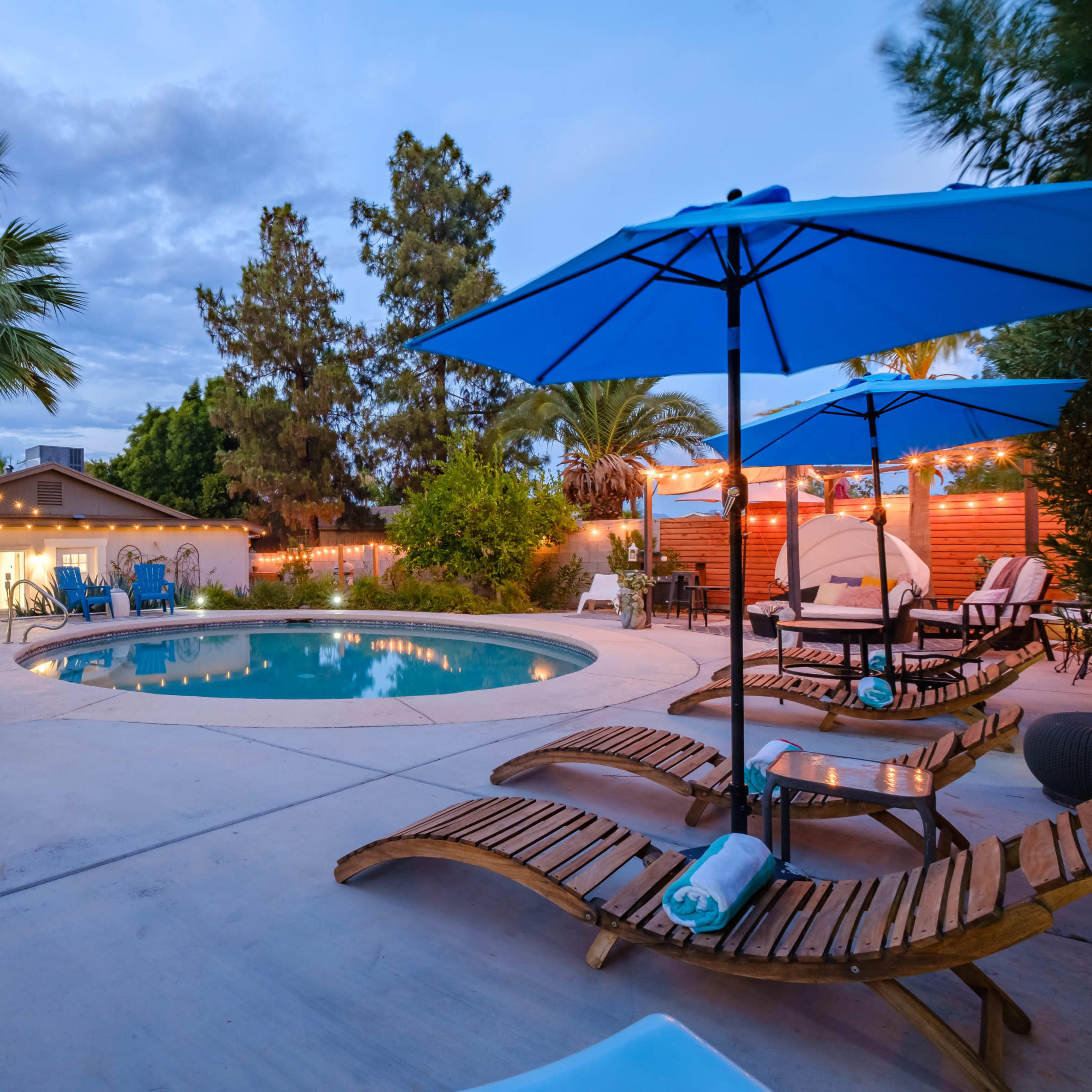 A spacious backyard features a swimming pool surrounded by lounge chairs and large umbrellas, with palm trees and string lights enhancing the outdoor setting.