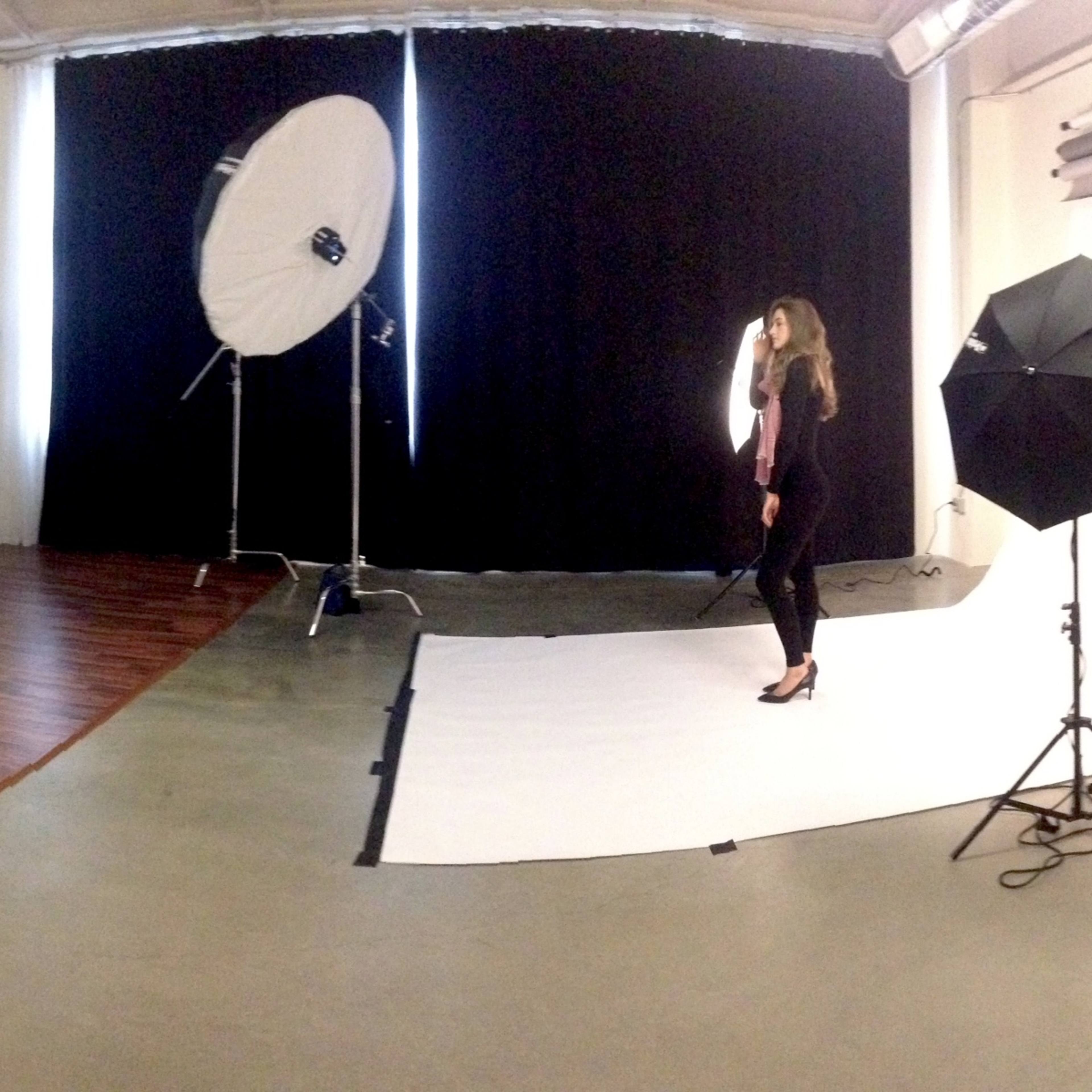 A model poses on a backdrop in a photography studio equipped with lighting and various props, while a few people observe from a distance.