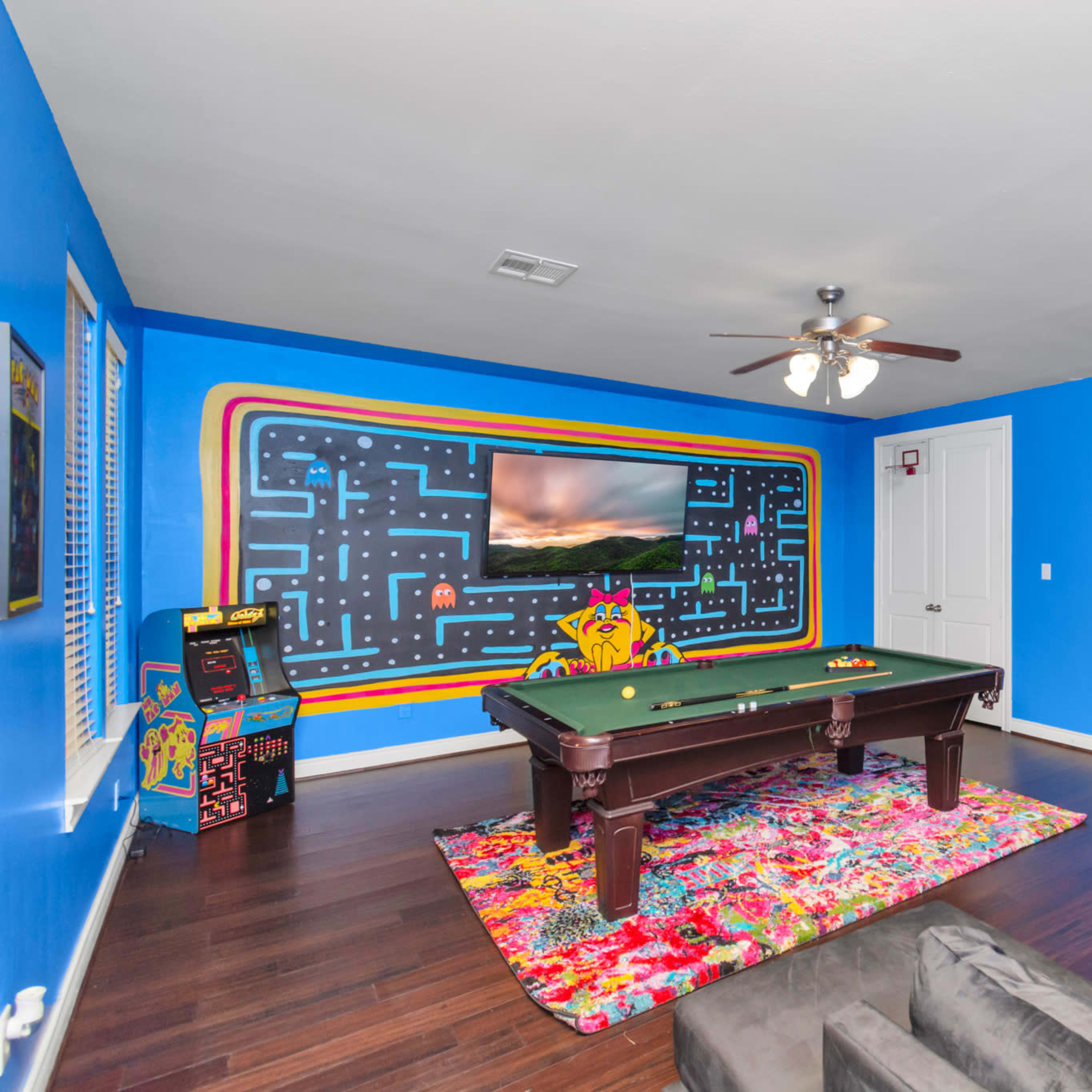 The room features a pool table in front of a colorful wall with a Pac-Man mural and an entertainment setup.