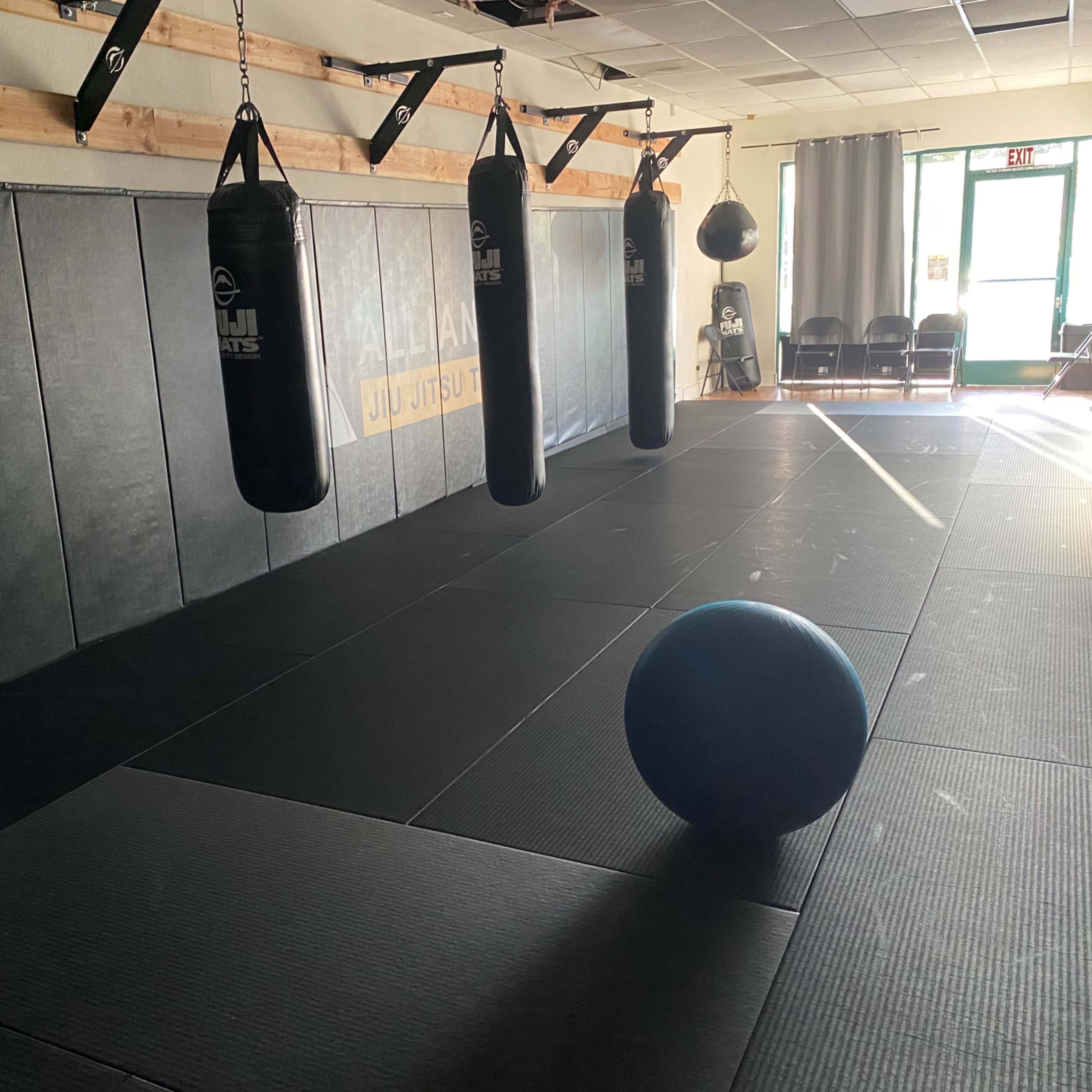 A sparsely furnished martial arts gym with black mats and hanging punching bags, featuring a large blue exercise ball positioned on the floor.