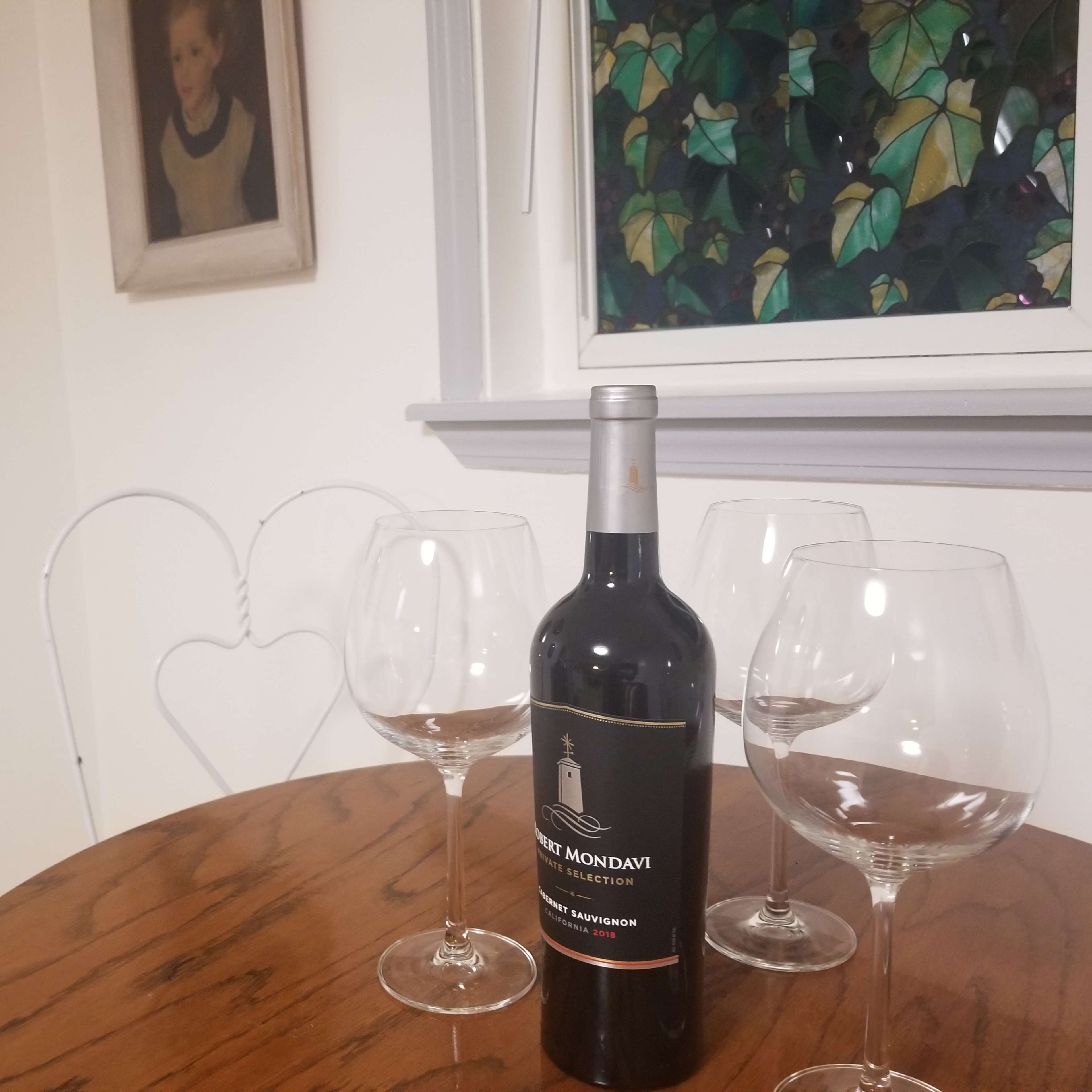 A bottle of wine and three wine glasses are placed on a wooden table in a room with two framed artworks and a window showing green leaves.