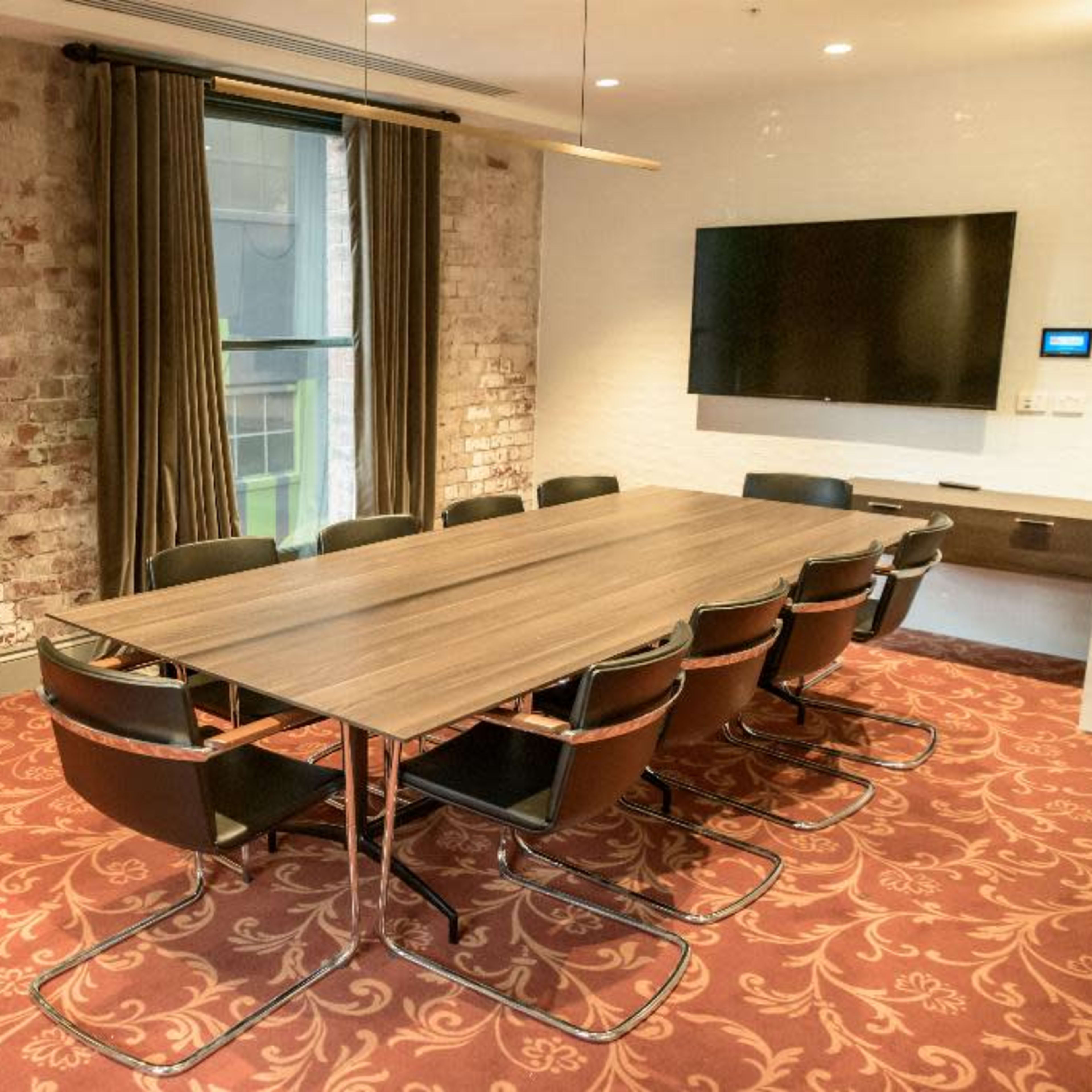 A conference room features a long wooden table surrounded by black chairs, with exposed brick walls and a wall-mounted television.