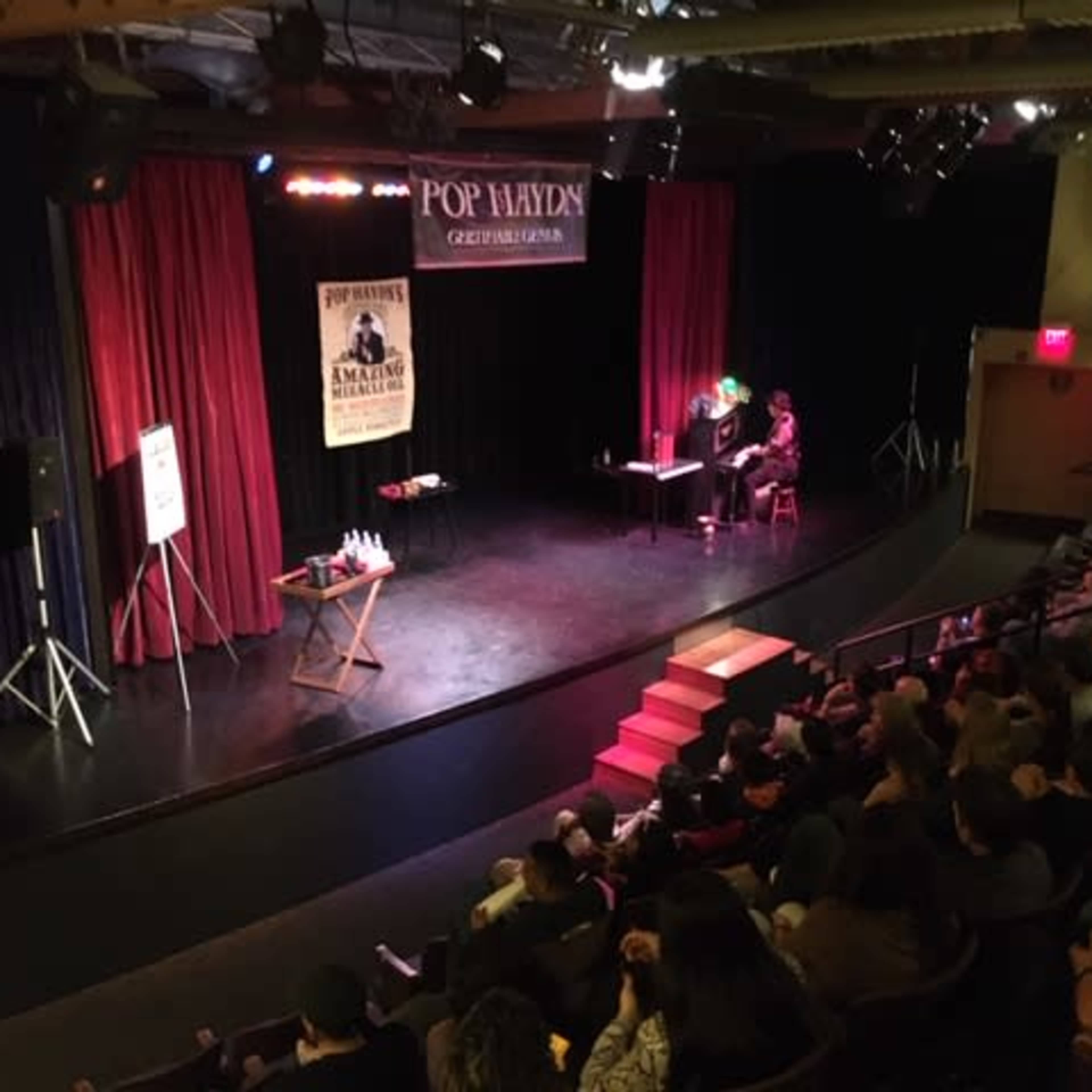 A stage is set for a performance, with two individuals seated at a table and an audience in the dimly lit theater.