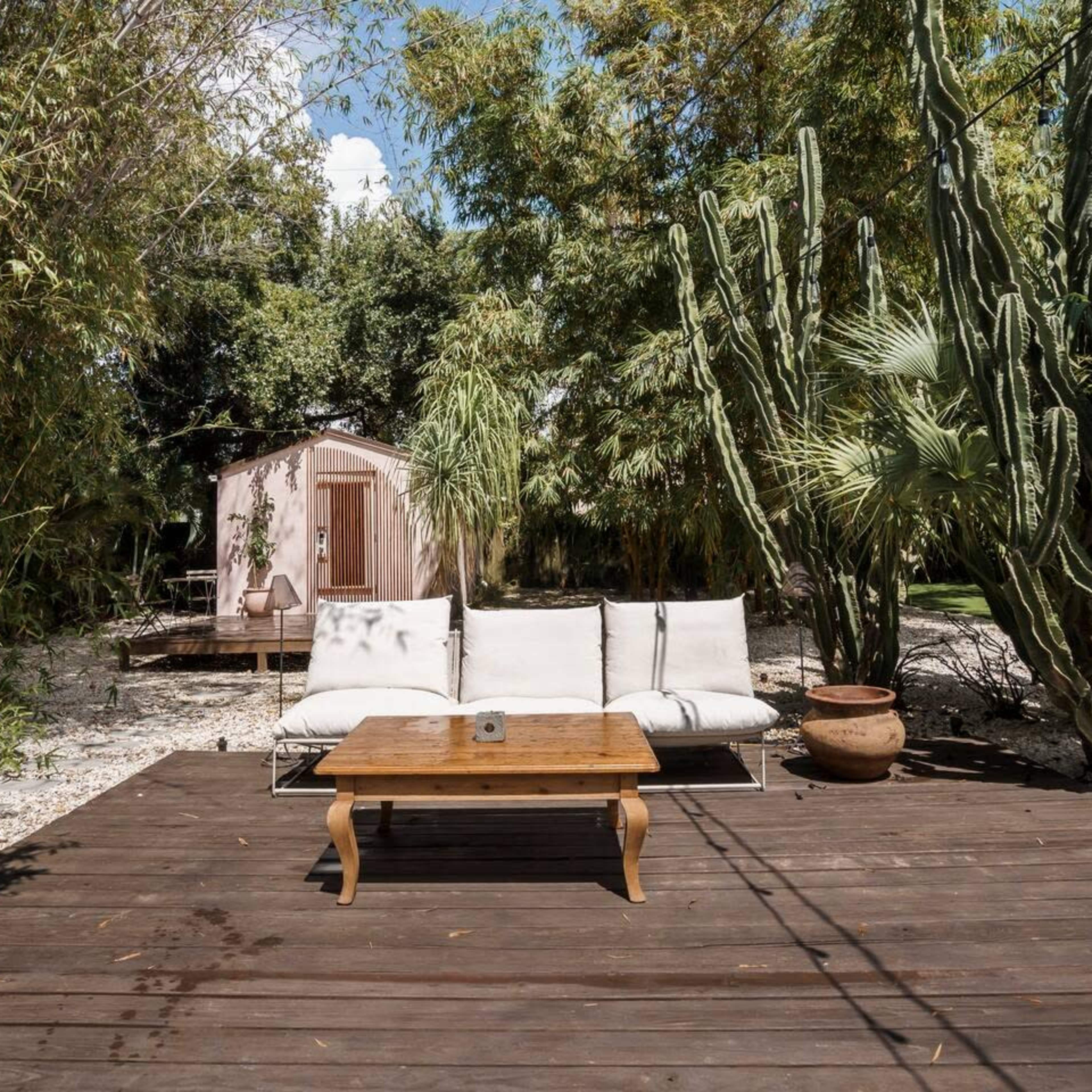 A wooden deck features a light-colored couch and table, surrounded by lush greenery and cacti, with a small cabin visible in the background.