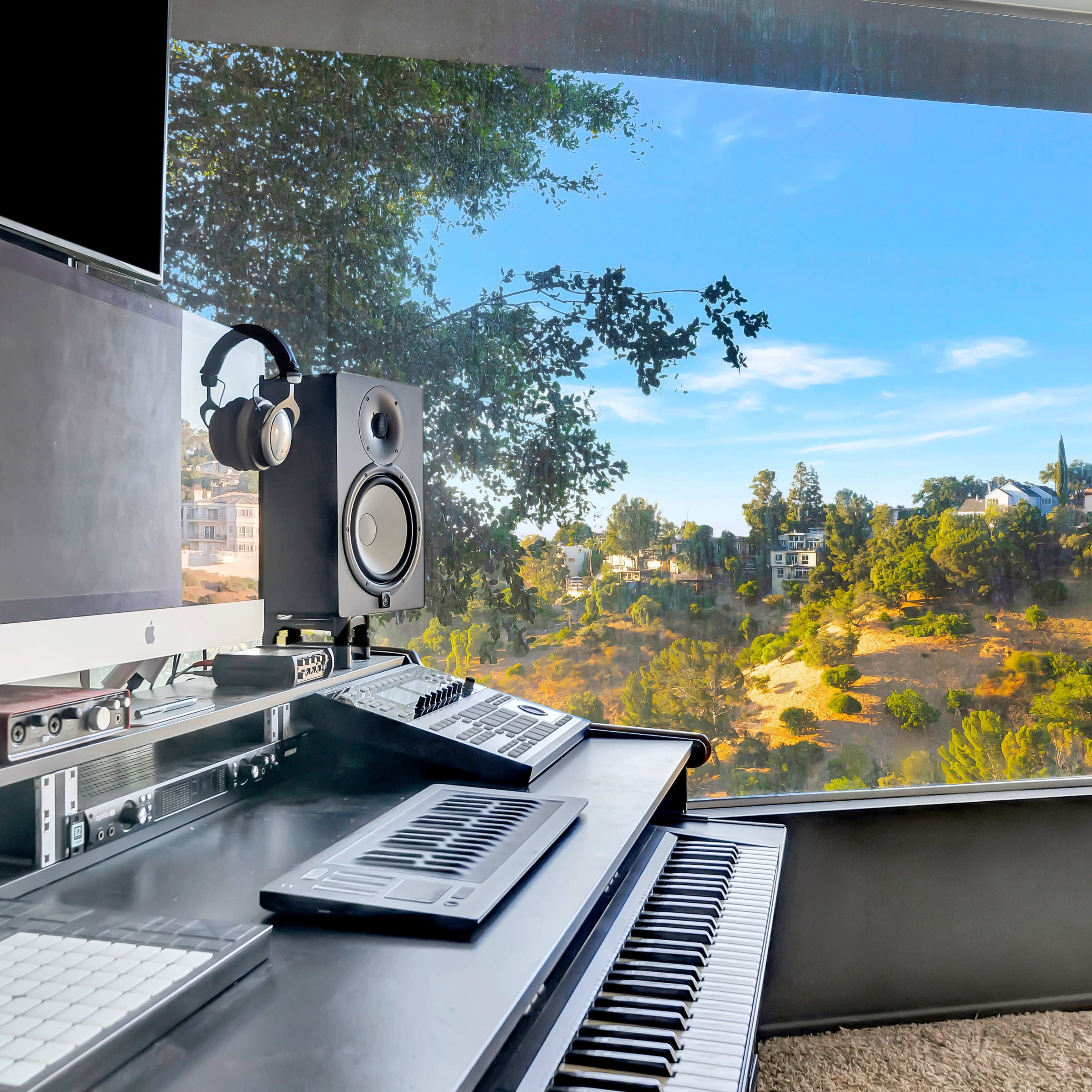 A music production studio features a digital audio workstation with speakers and headphones, overlooking a scenic view of a landscape through large windows.