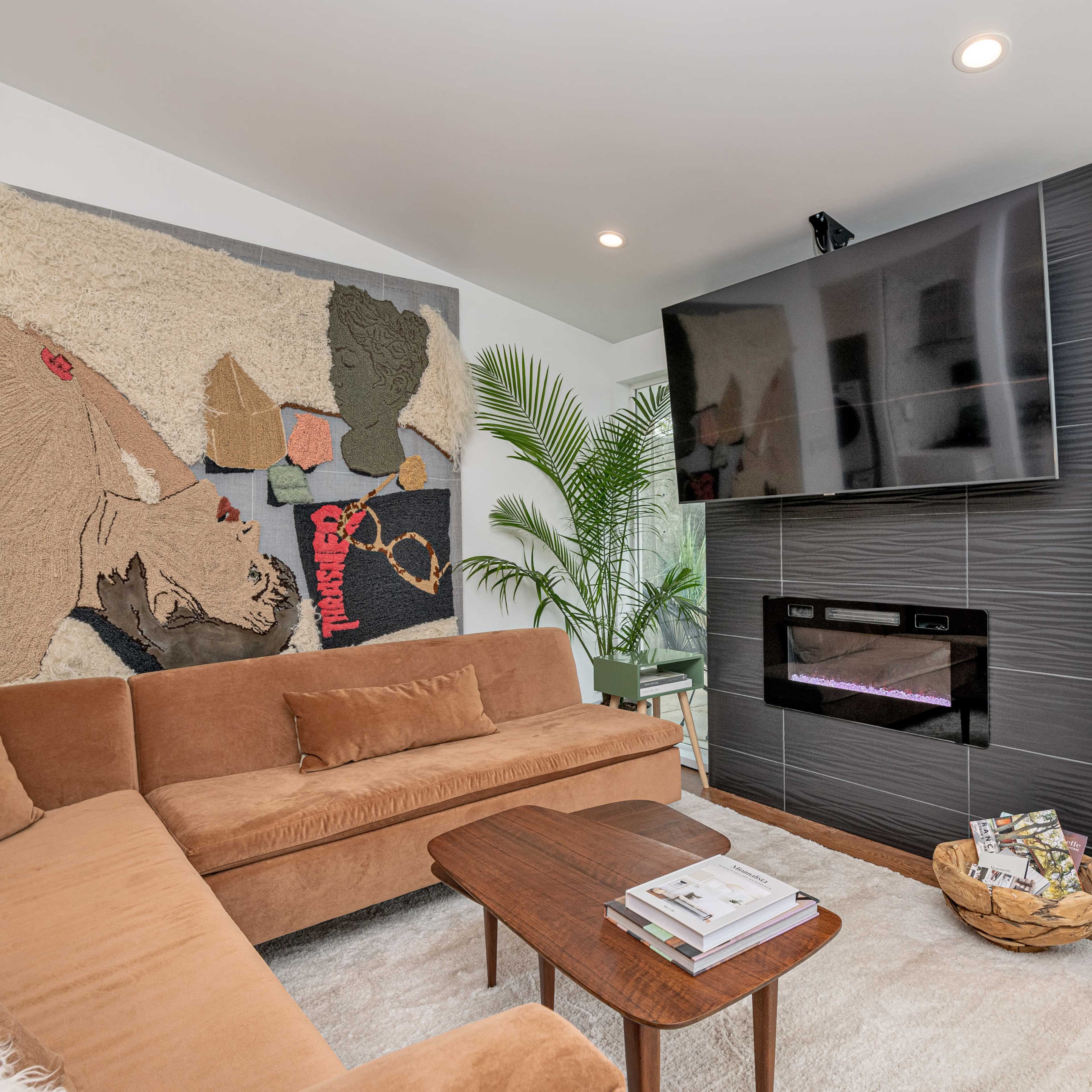 A modern living room featuring a brown sectional sofa, a wall-mounted television, and a decorative tapestry above a sleek electric fireplace.