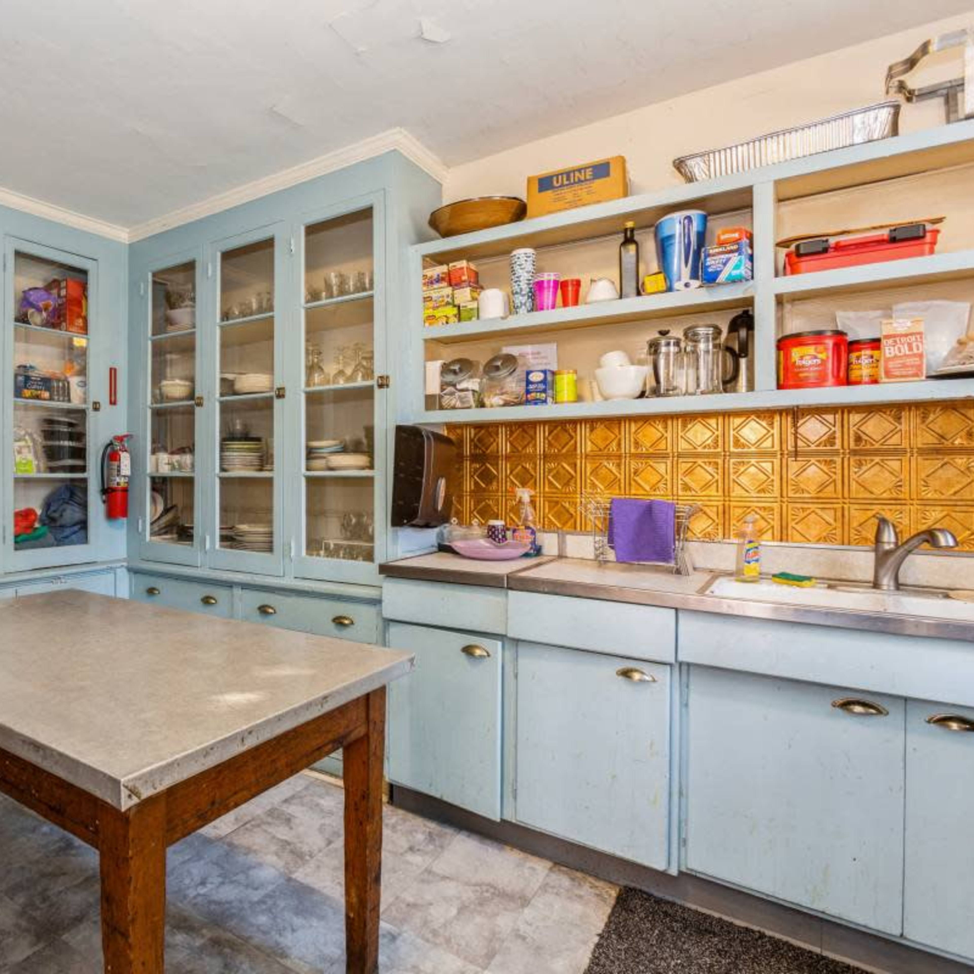 A kitchen with light blue cabinetry, a central table, and shelves filled with various dishes and food items.