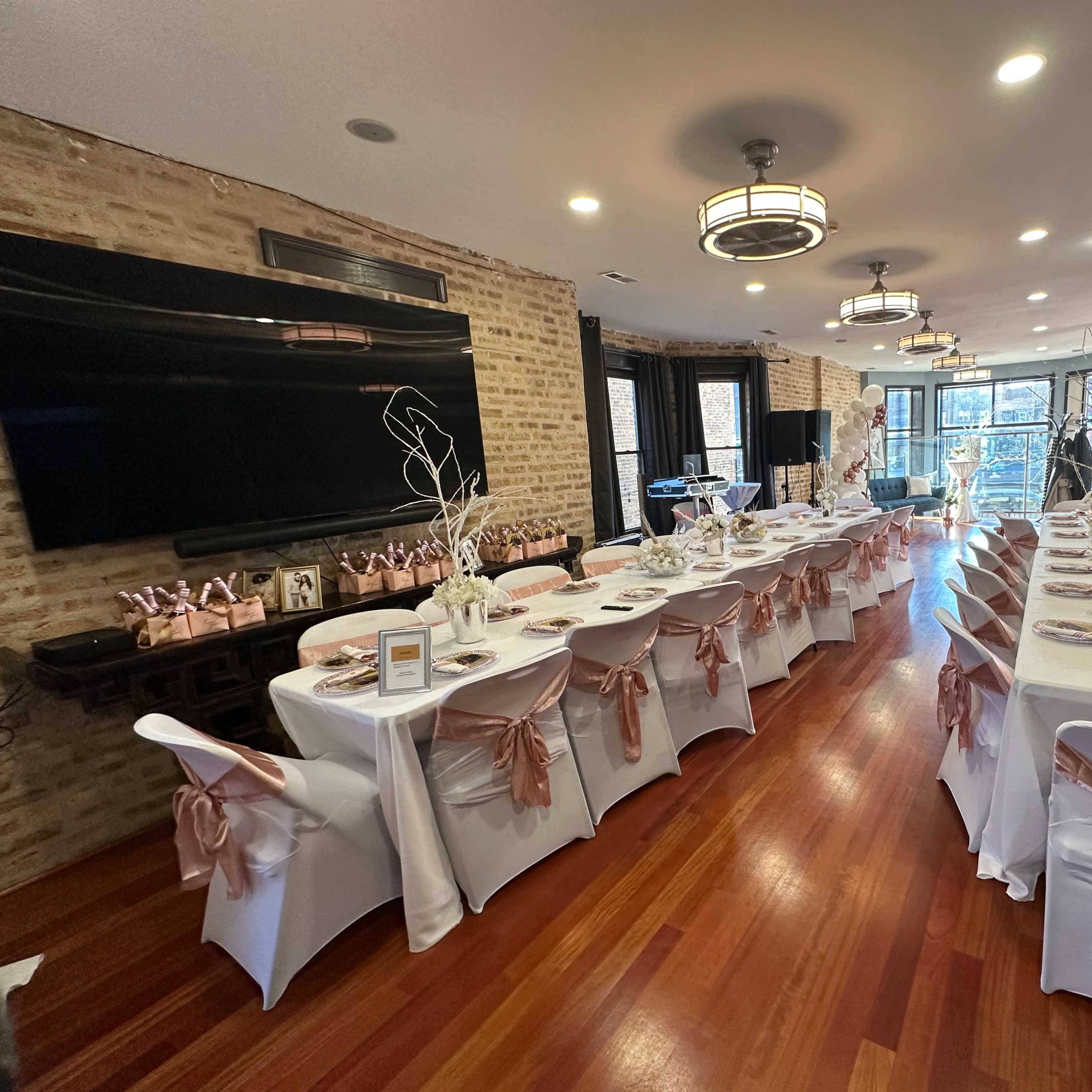 A decorated event space features a long dining table set for a celebration, with white tablecloths and pink satin bows on the chairs.