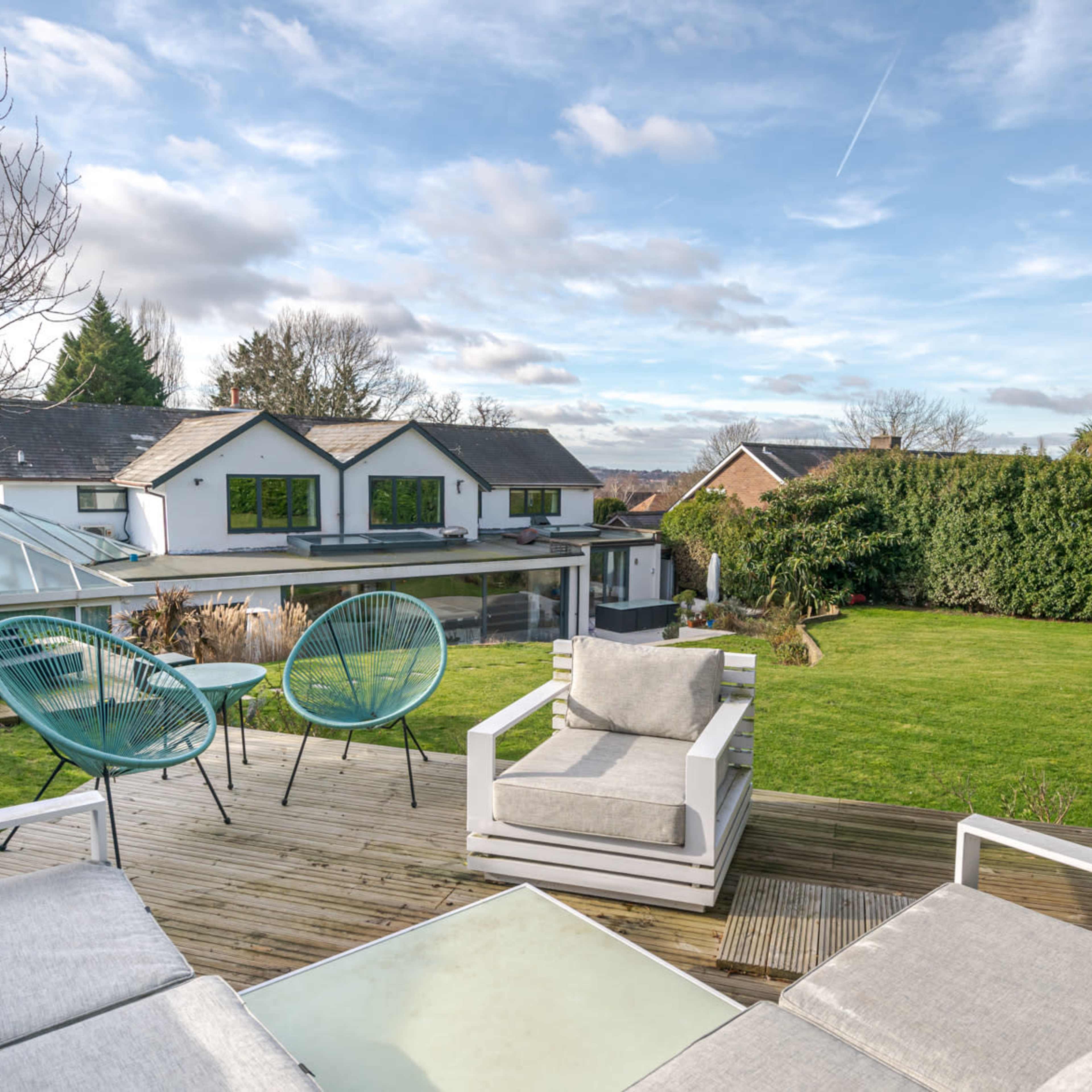 A wooden deck overlooks a landscaped garden and two houses, with modern outdoor furniture positioned for relaxation.