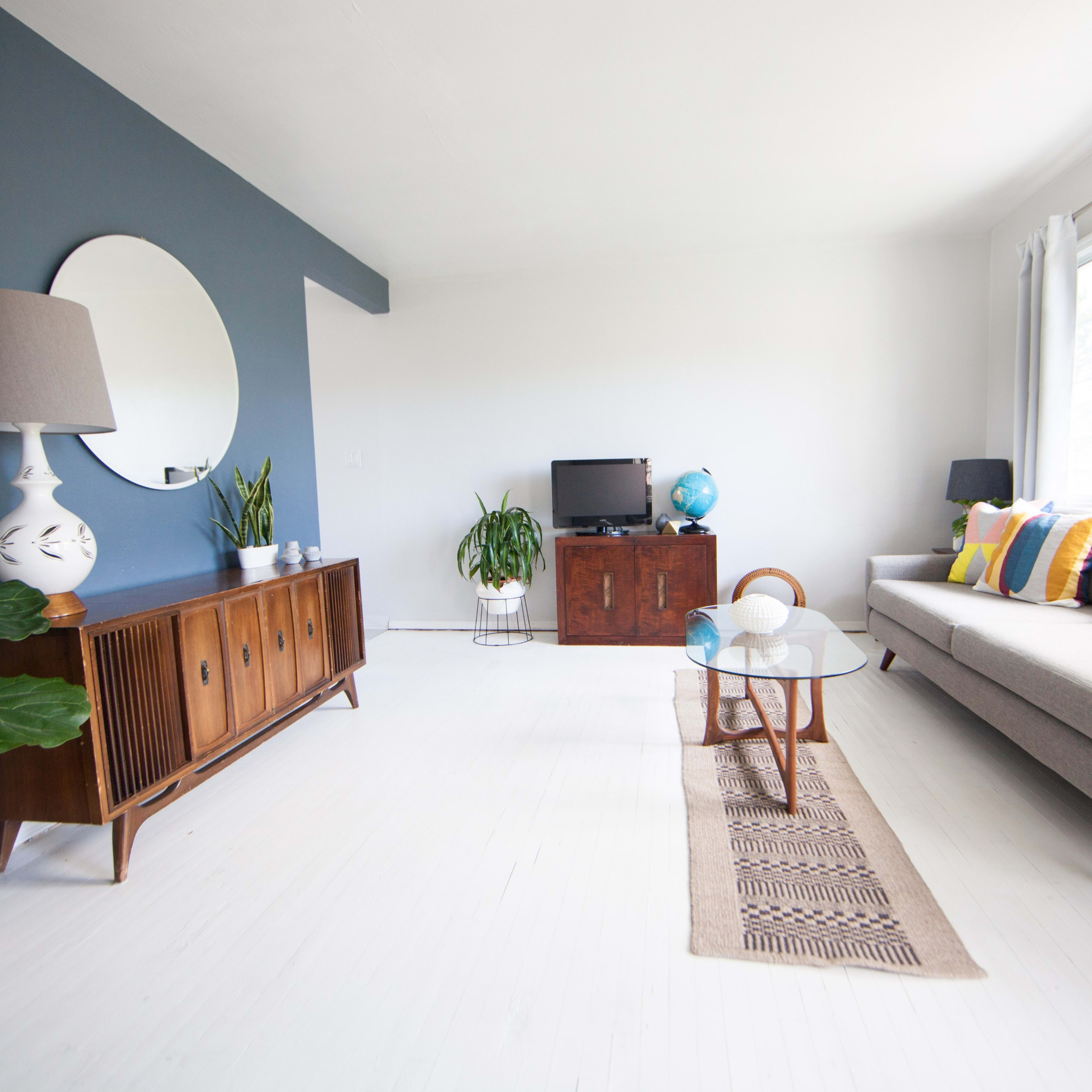 The living room features a gray sofa, a wooden coffee table, and a media console against a two-tone wall, with a large window letting in natural light.