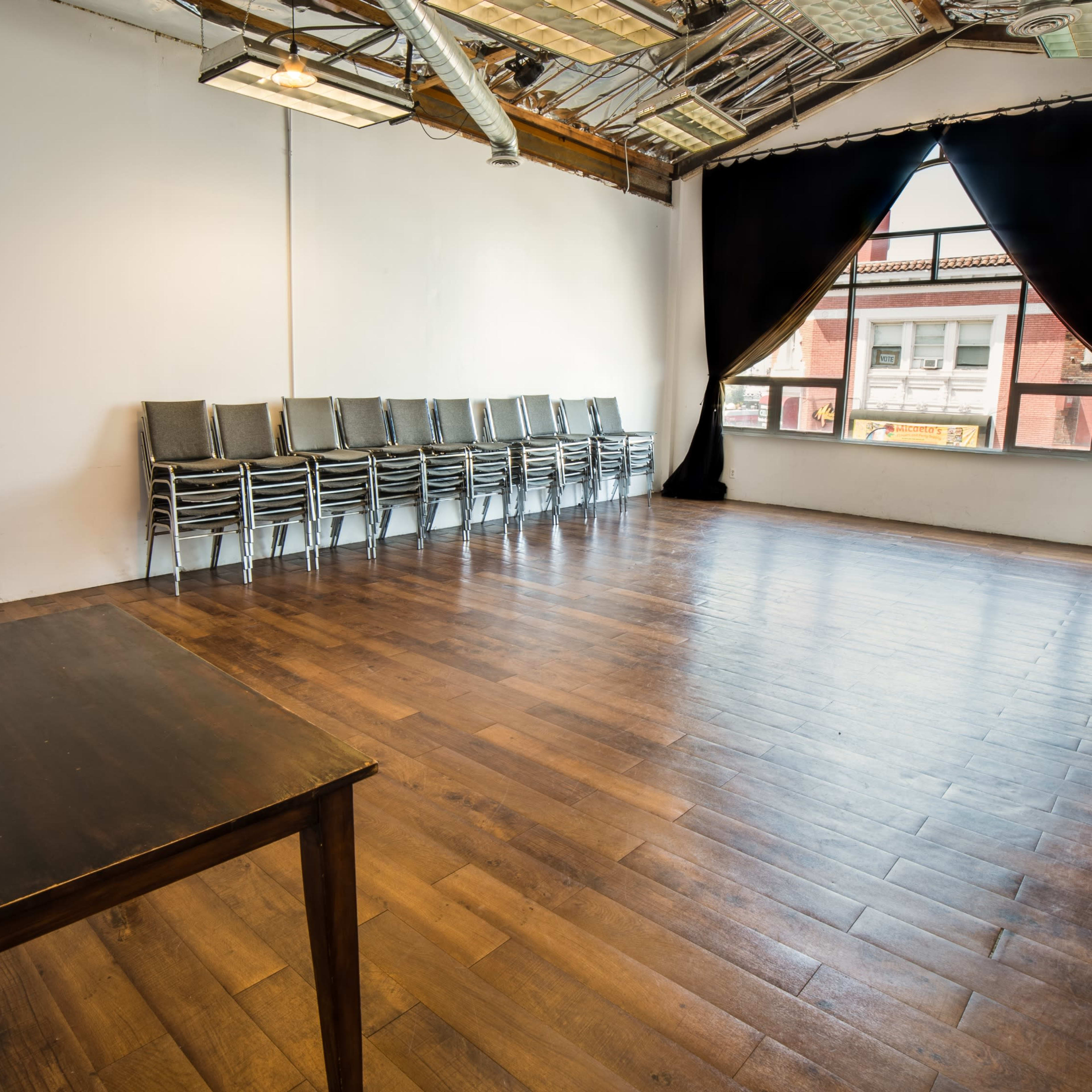 An empty room with hardwood floors, a large window, black curtains, a stack of chairs against the wall, and a table with equipment.