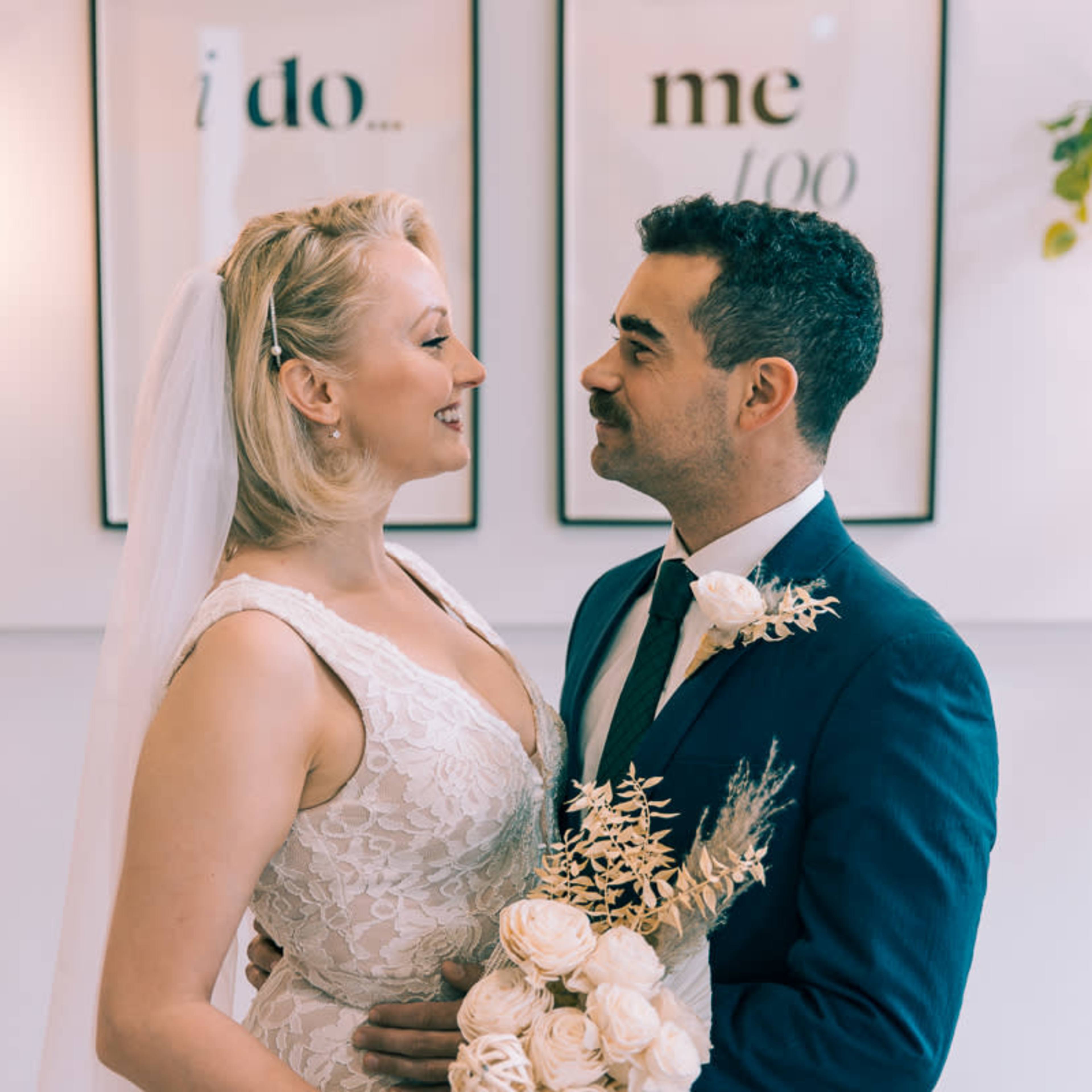 A bride and groom stand close together, smiling at each other while holding a bouquet in a decorated indoor setting.