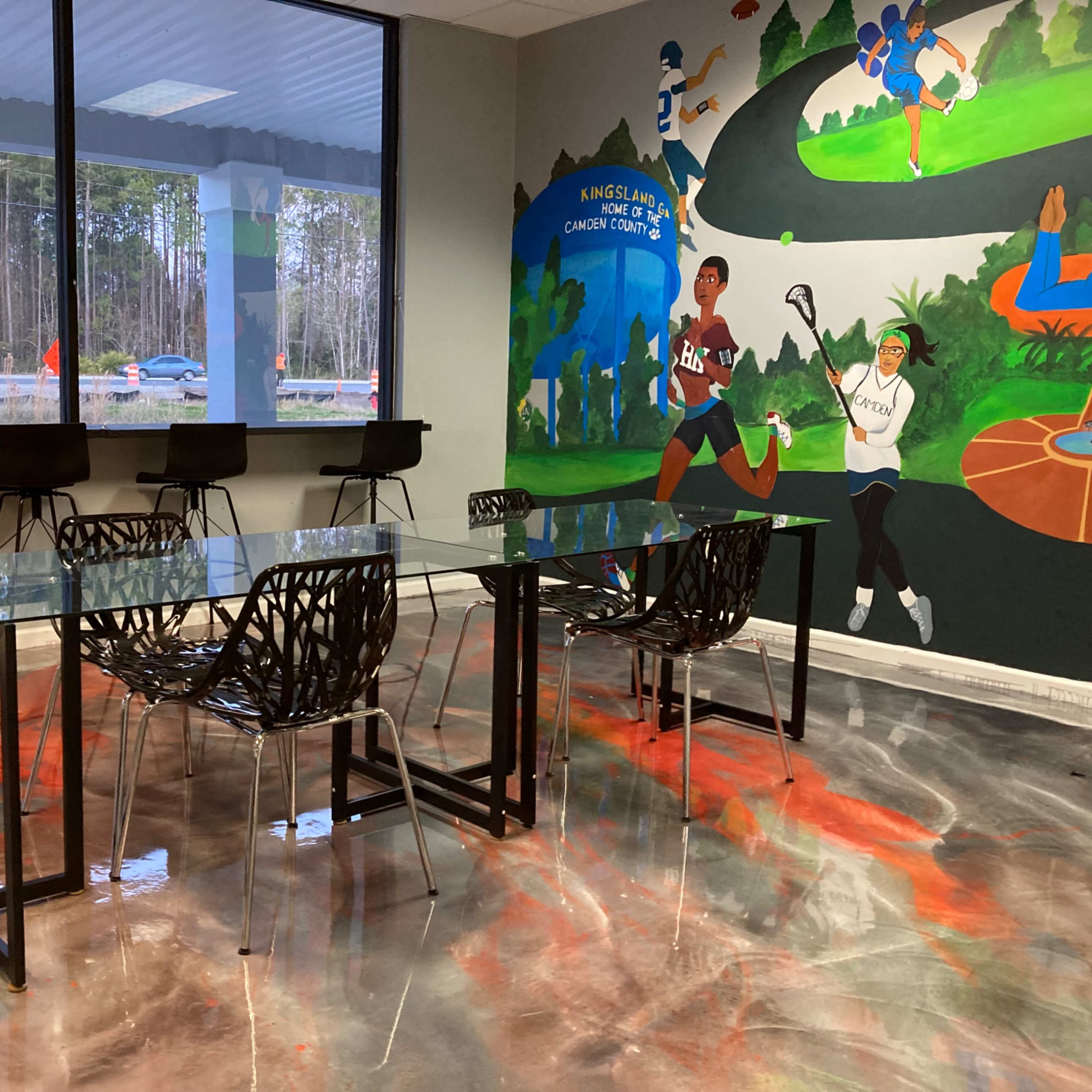 A modern workspace features a glass table surrounded by black chairs and a colorful mural on the wall.
