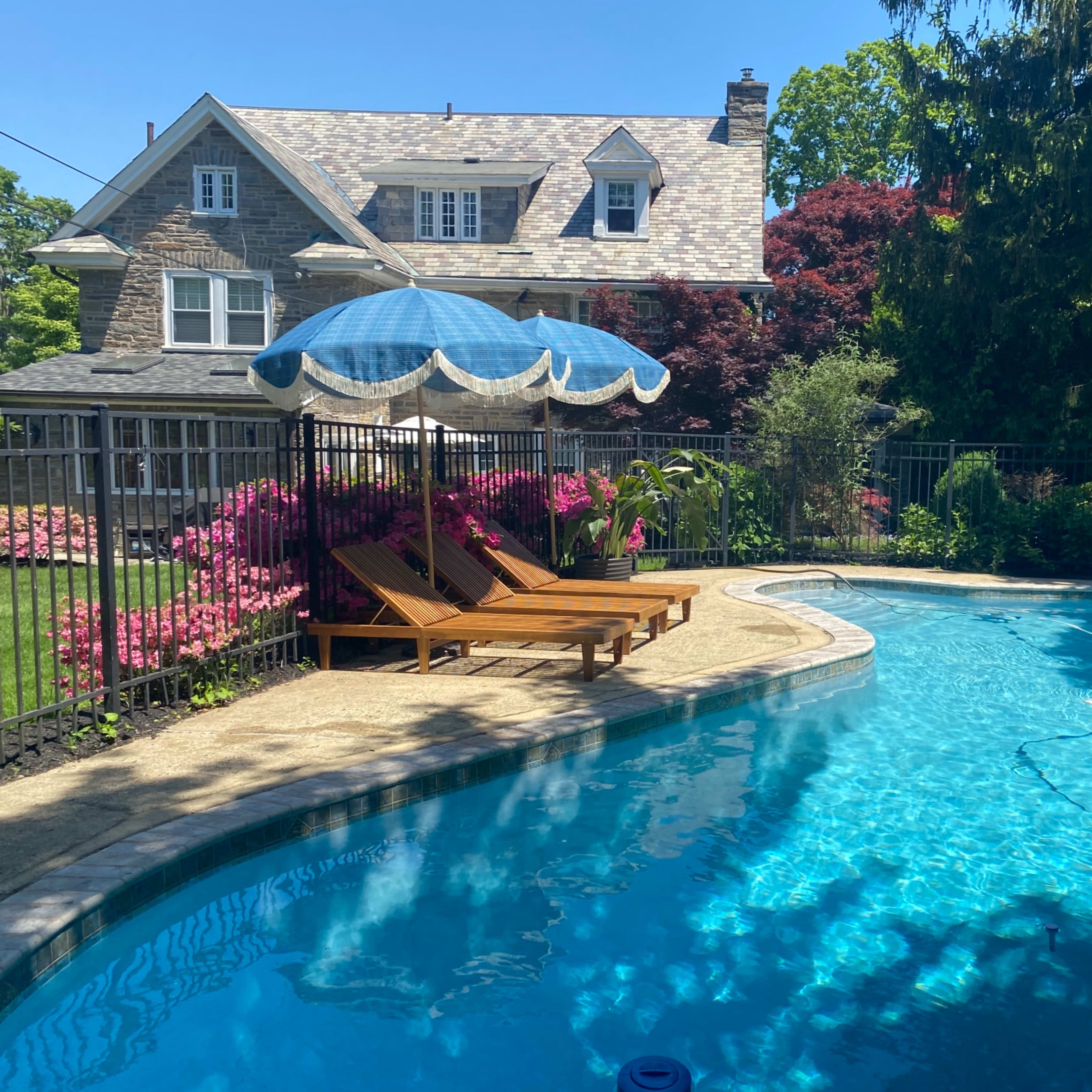 A swimming pool with clear blue water is surrounded by a fence, deck chairs, and vibrant flowers beneath a blue umbrella.