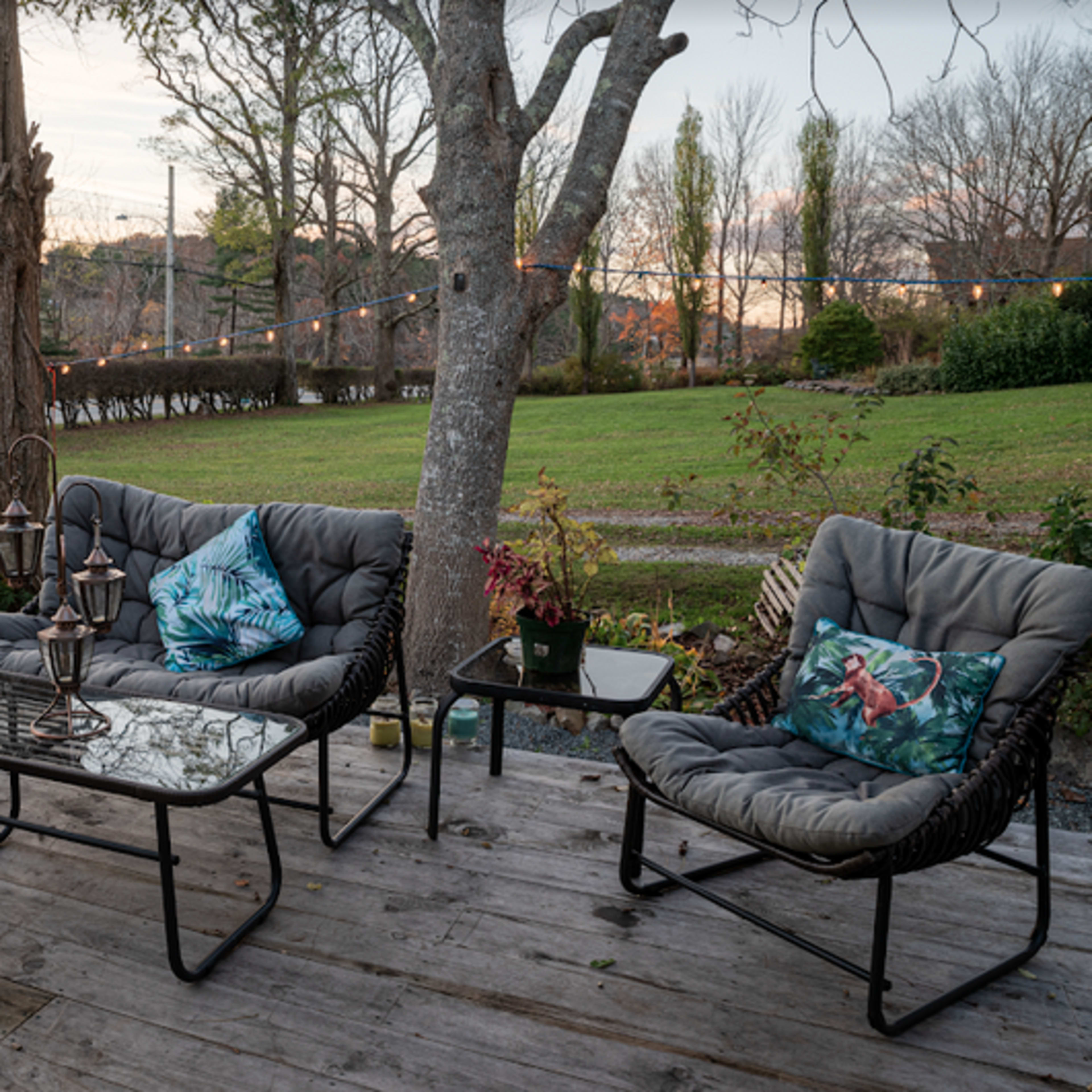 A patio features a seating arrangement with gray cushions and tropical-themed pillows, alongside a glass table, set against a backdrop of trees and distant greenery at sunset.