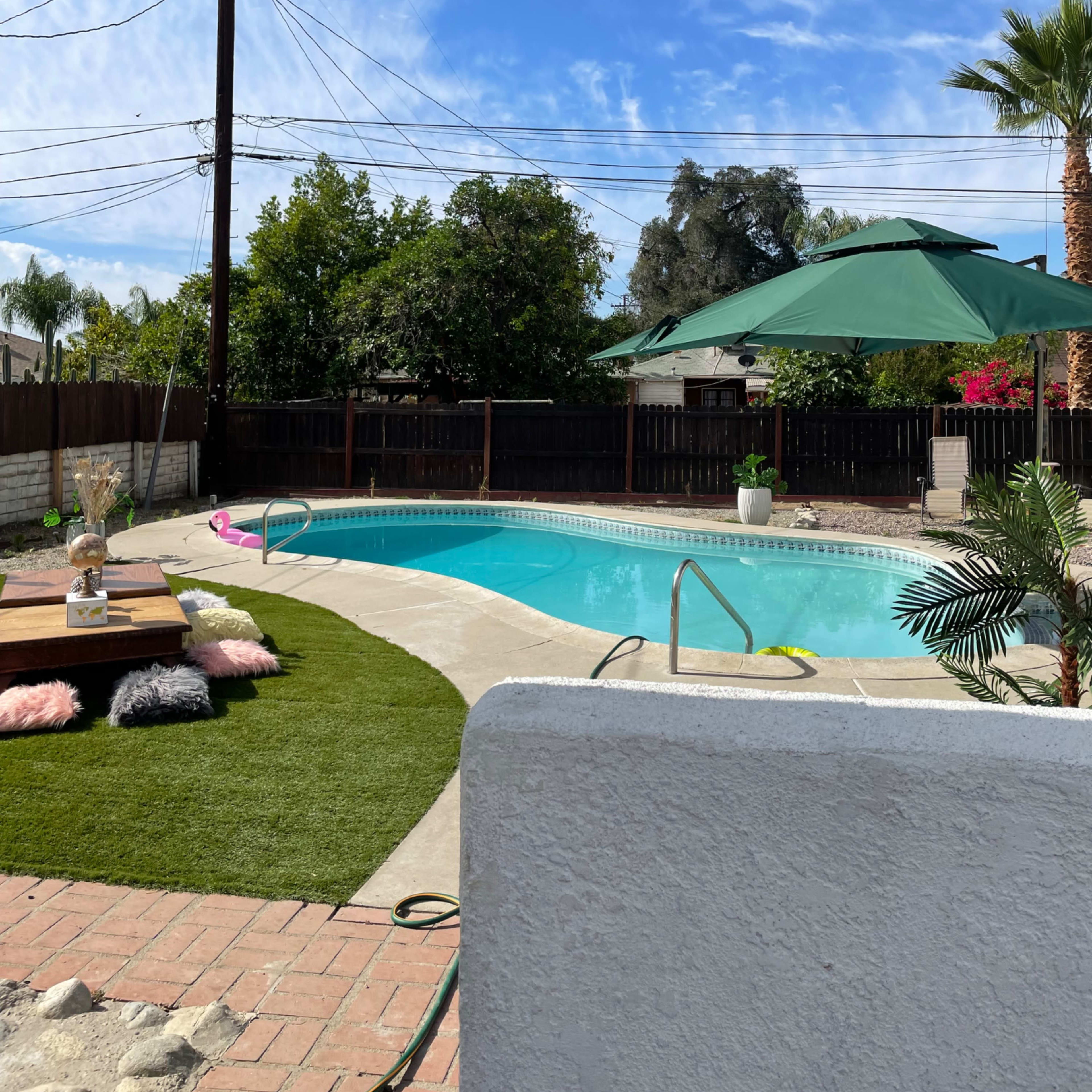 A swimming pool surrounded by green grass, a wooden deck with seating, and palm trees, under a clear blue sky with power lines in the background.