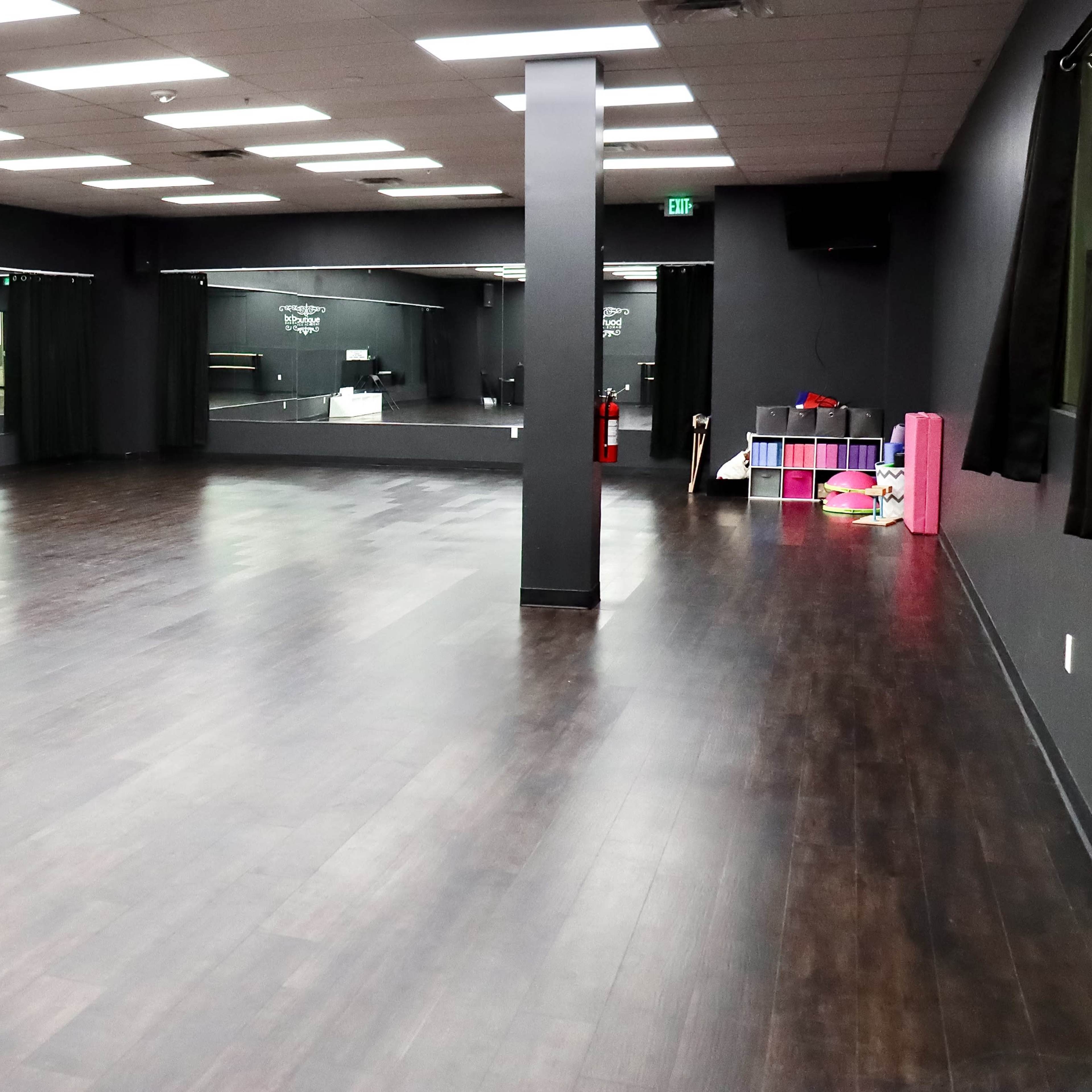 A spacious dance studio with dark wood flooring, black walls, and large mirrors on one side.