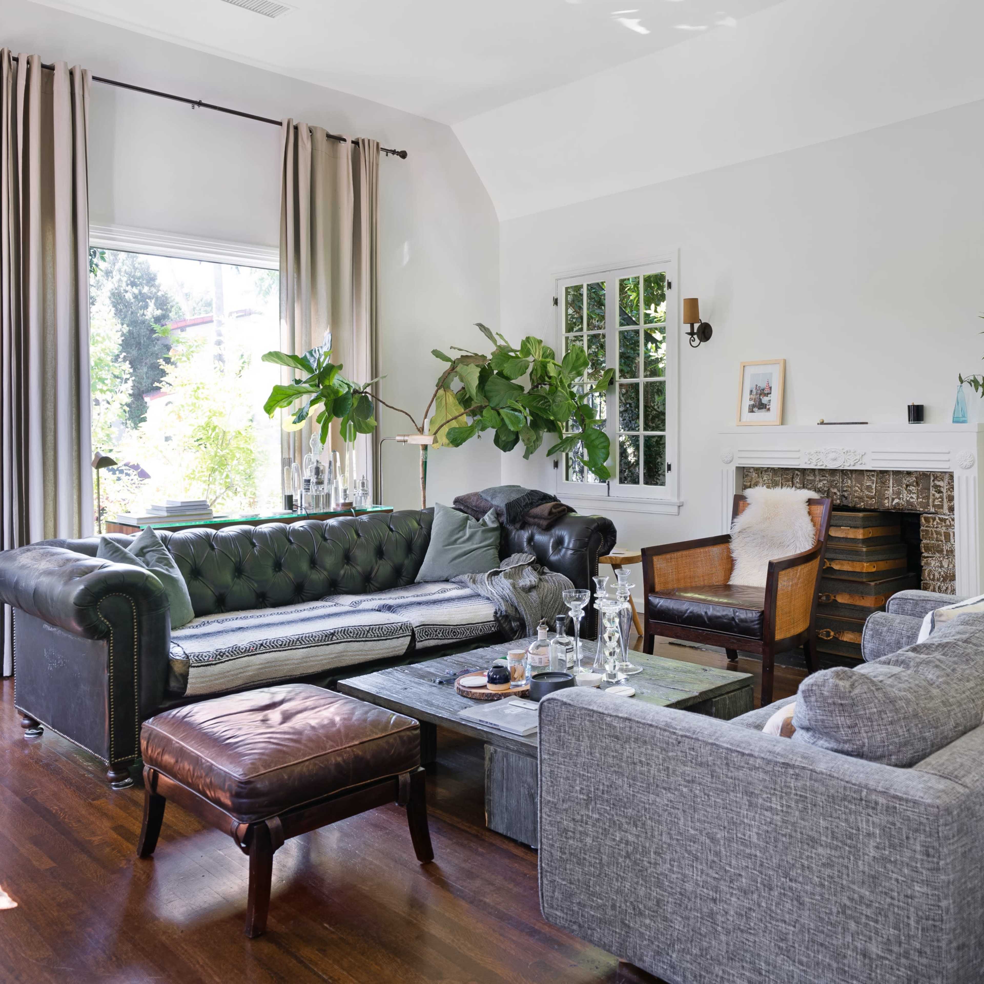 The living room features a mix of a dark green chesterfield sofa, a gray sectional, and a wooden armchair, arranged around a coffee table, with abundant natural light from large windows.