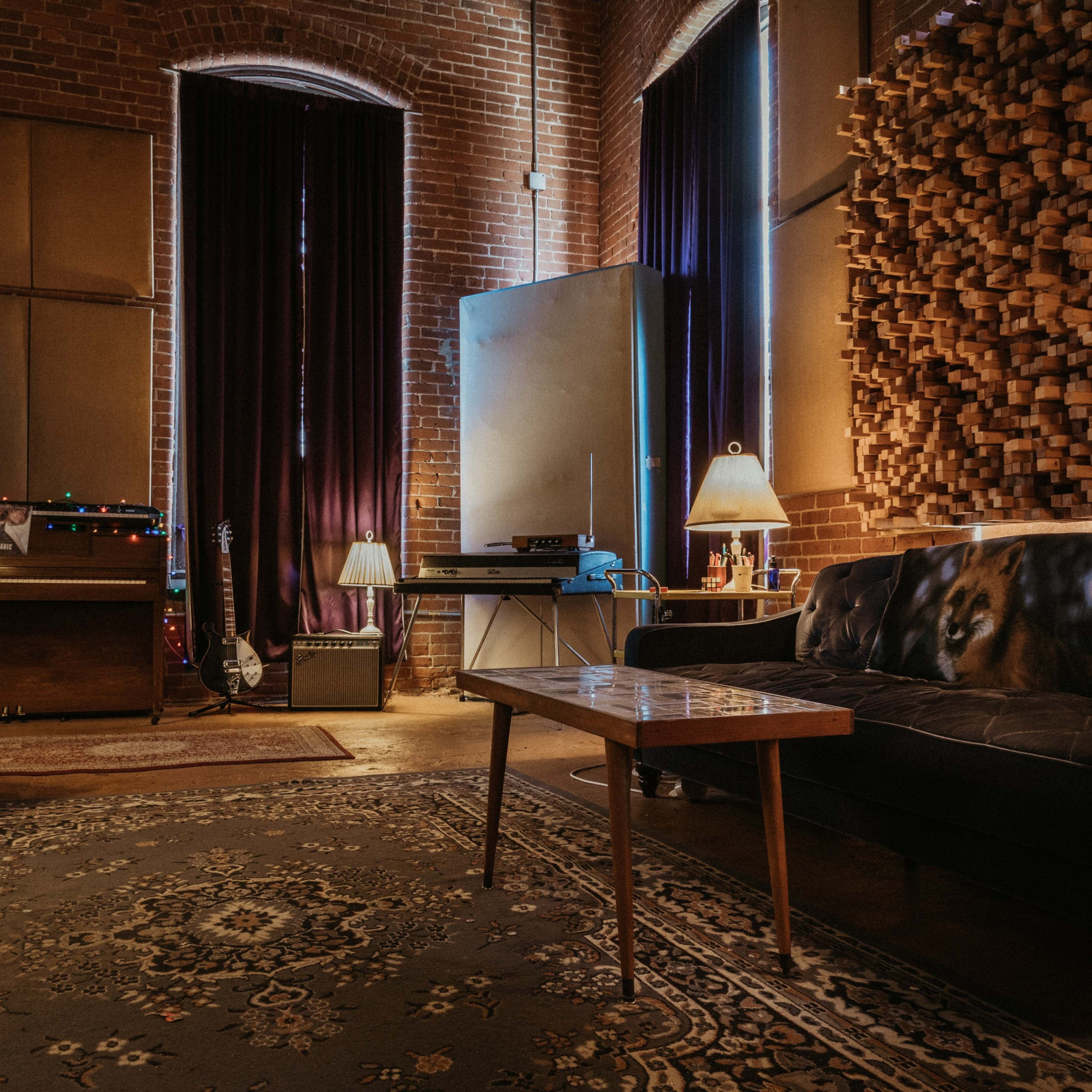 A cozy music studio features a black couch, wooden sound paneling, a vintage recording console, and soft lighting from two lamps.