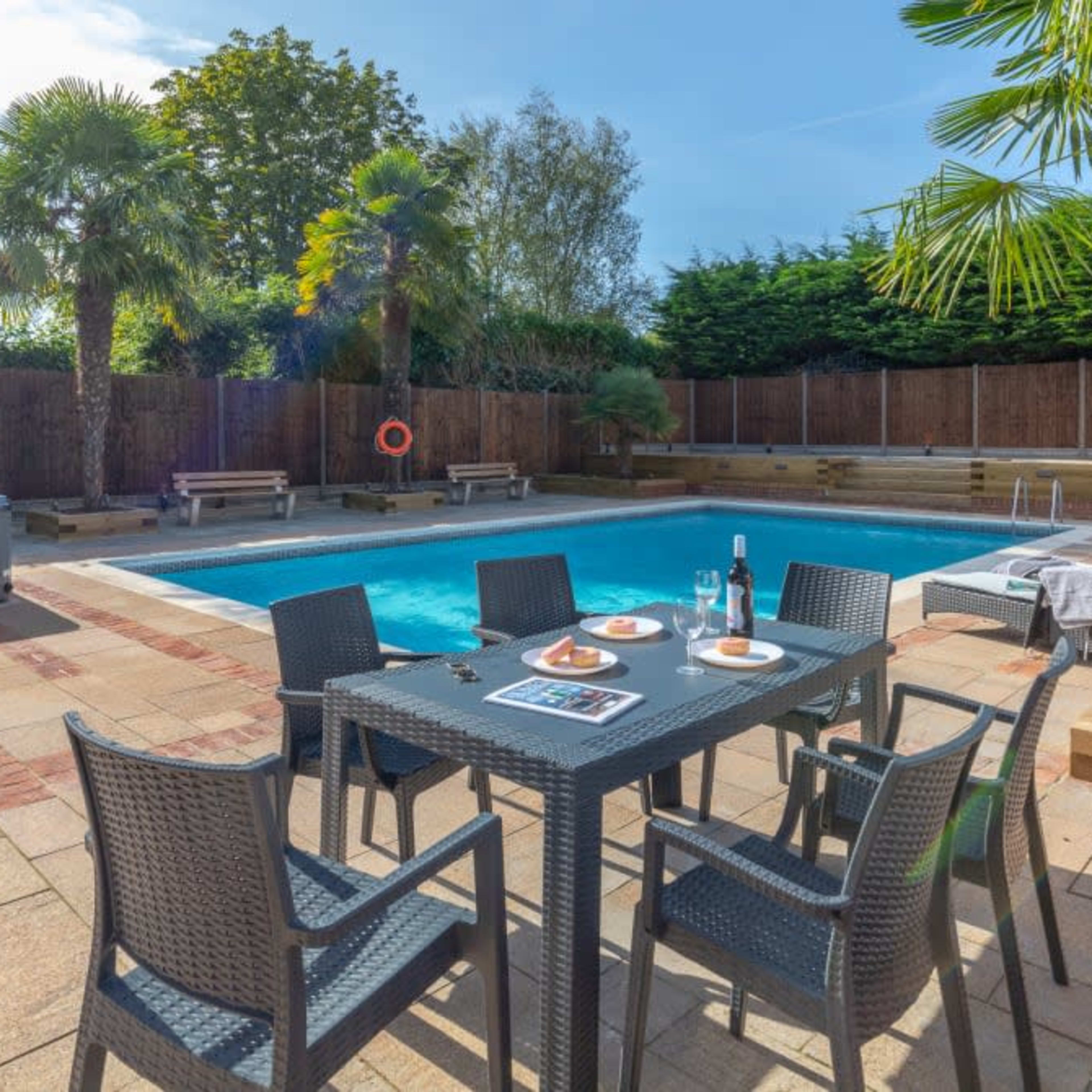 A backyard scene featuring a swimming pool surrounded by palm trees, lounge chairs, and a dining set with plates on a stone patio.