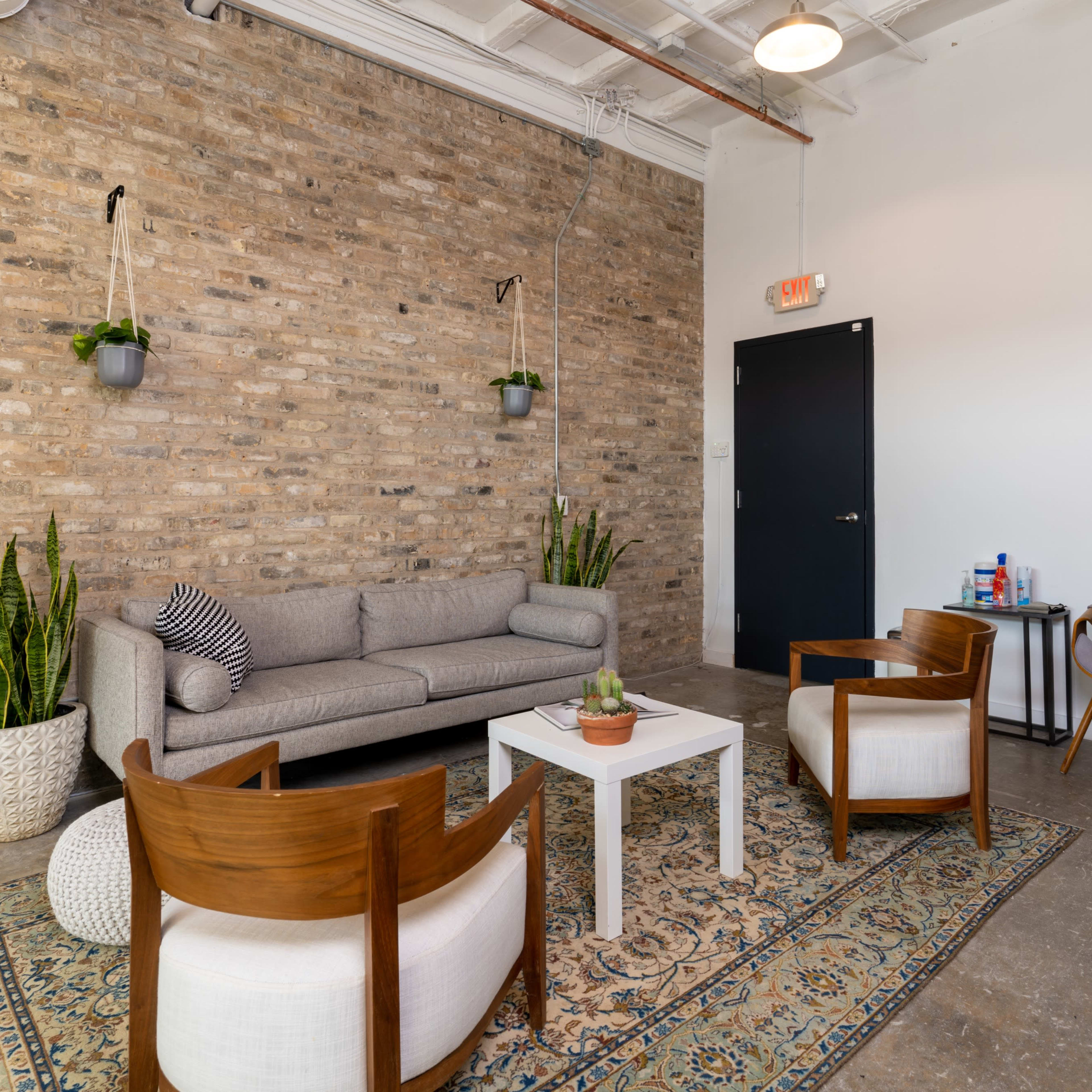 A seating area with a gray sofa, two wooden chairs, a coffee table, and a patterned rug against a brick wall with potted plants.