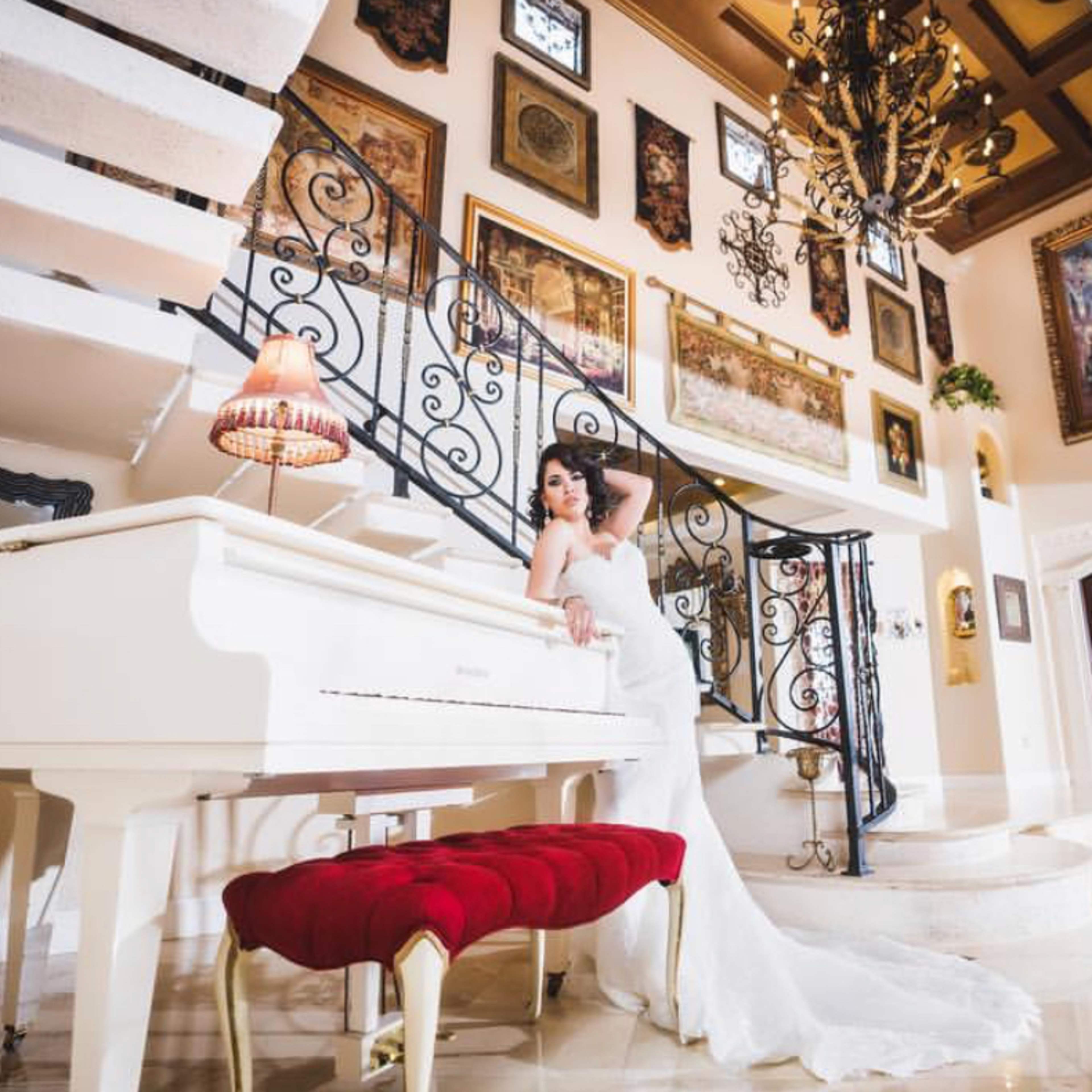 A woman in a white gown poses beside a grand white piano in a richly decorated room featuring ornate staircases and framed artwork on the walls.
