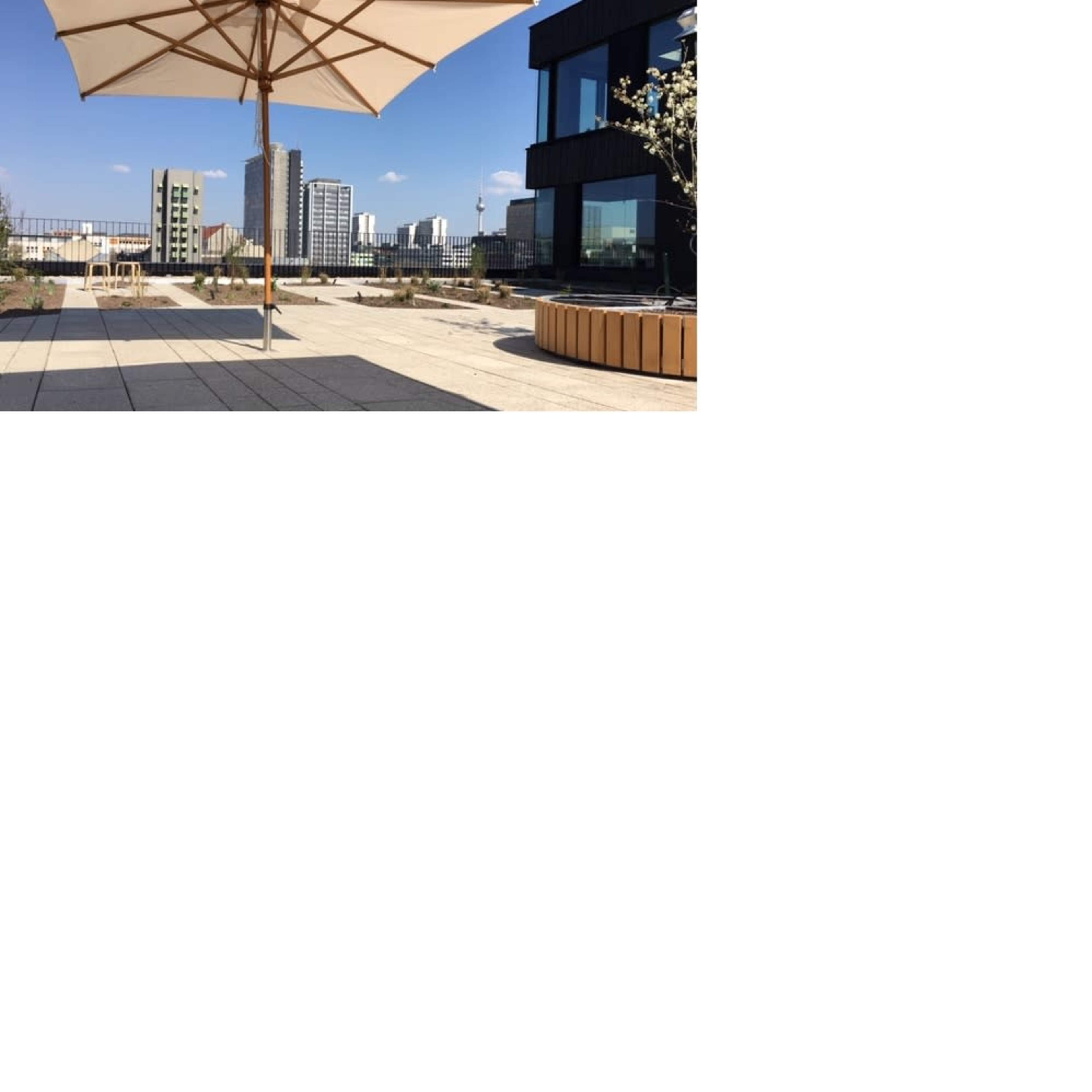 A large umbrella stands on a rooftop terrace overlooking modern buildings and a clear blue sky.