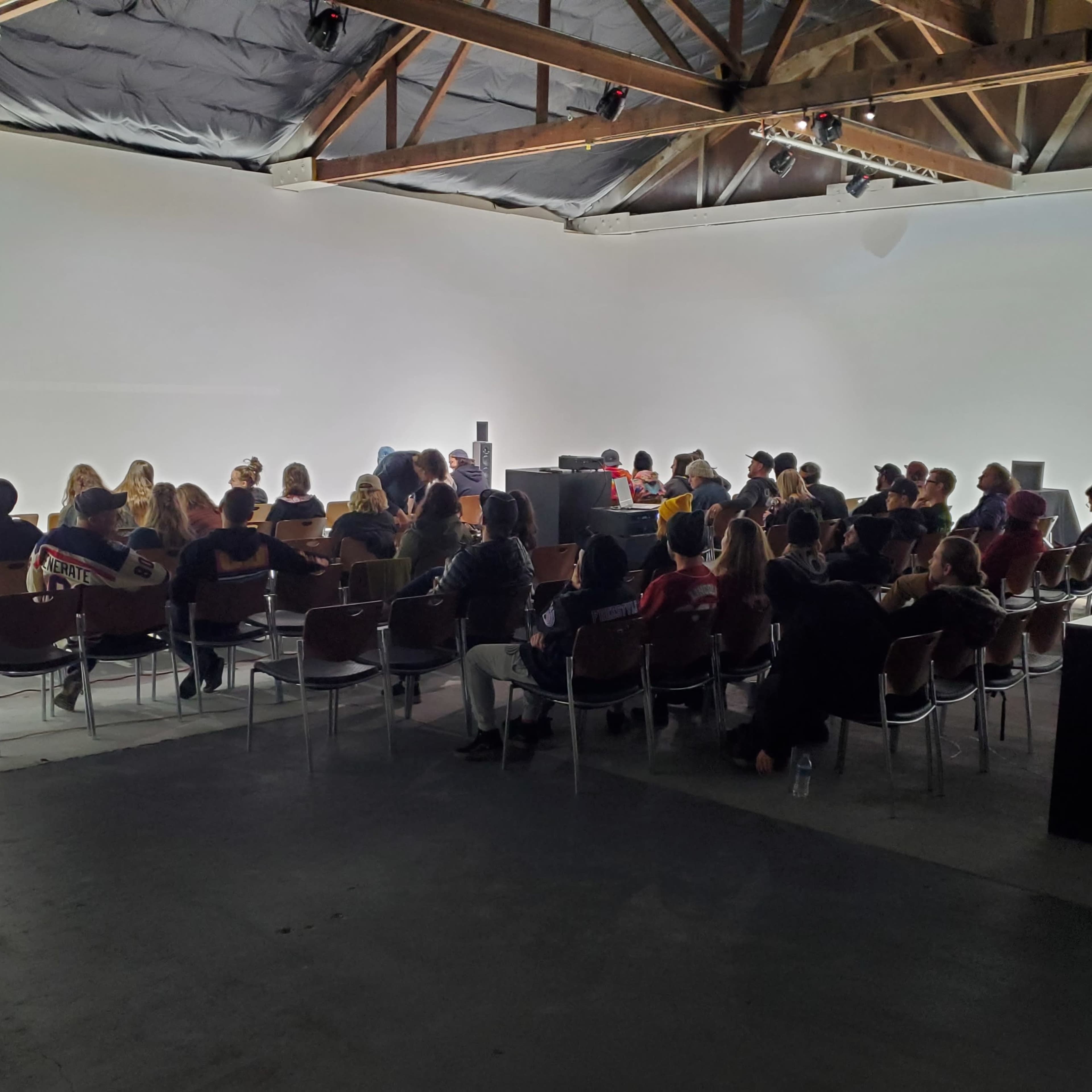A large group of people is seated in chairs facing a raised platform in a minimalistic, well-lit space with exposed wooden beams.