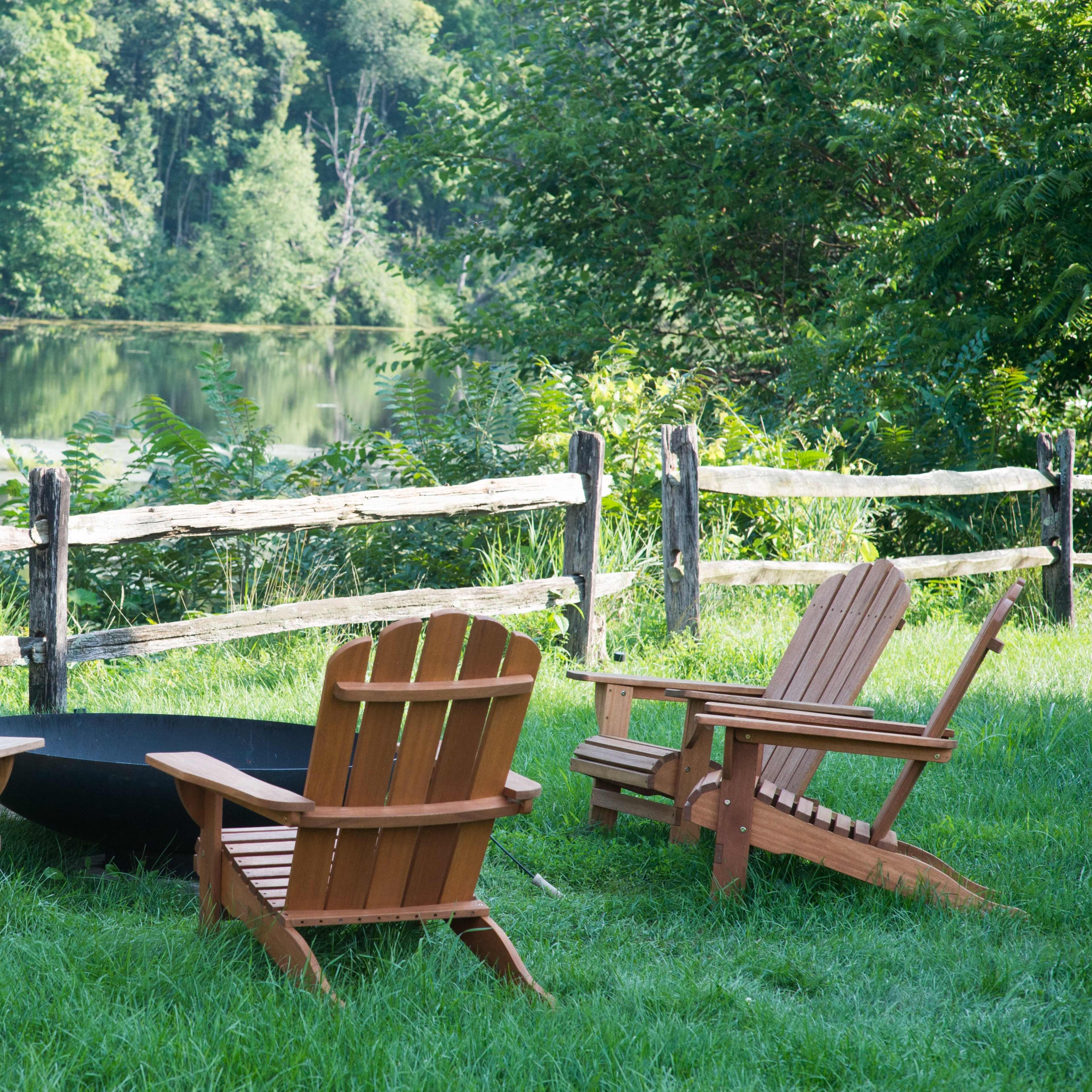 Three wooden Adirondack chairs are arranged around a black fire pit near a serene body of water, framed by a wooden fence and surrounded by greenery.