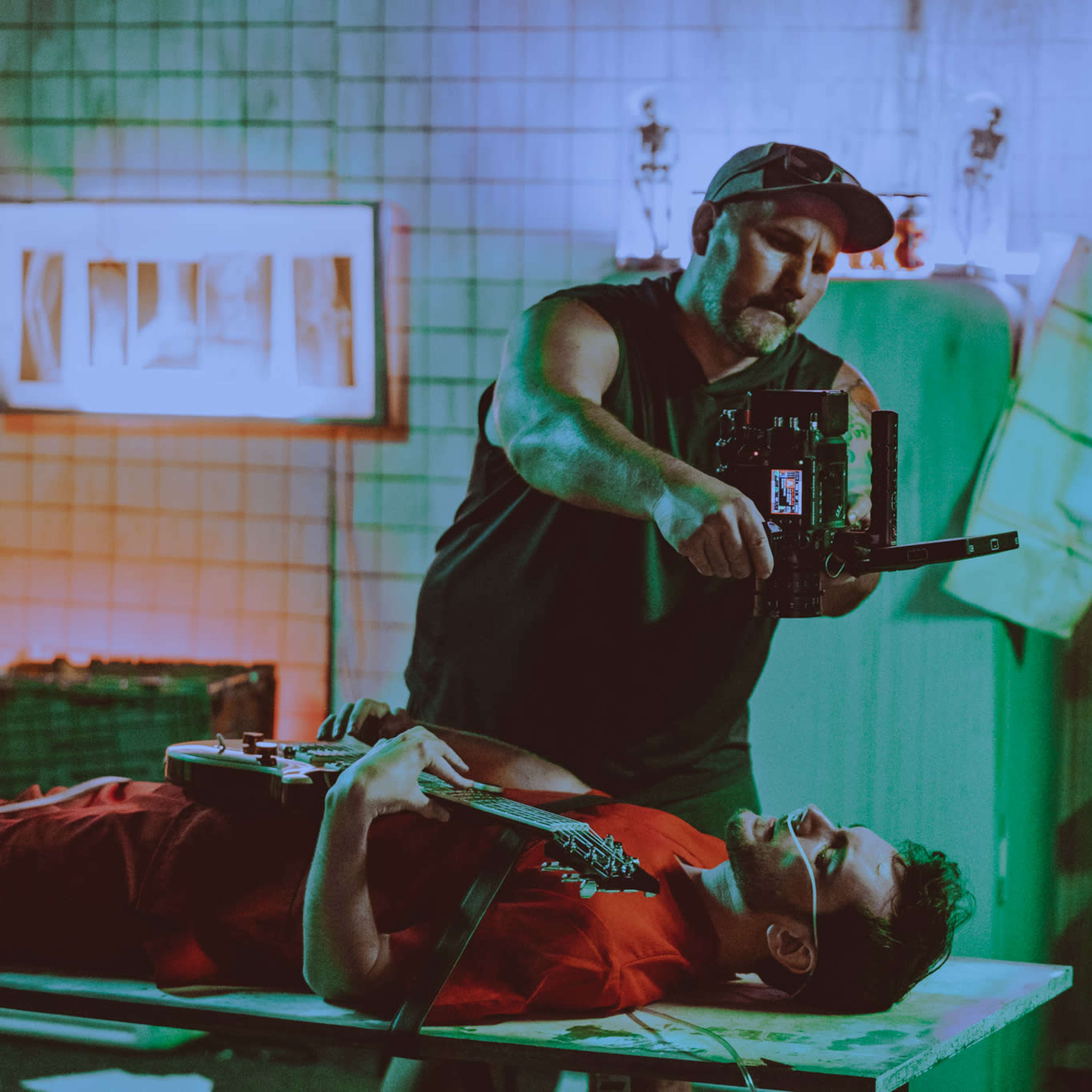 A man in a cap films another man lying on a table with a guitar in a dimly lit room featuring colored lighting and various props.