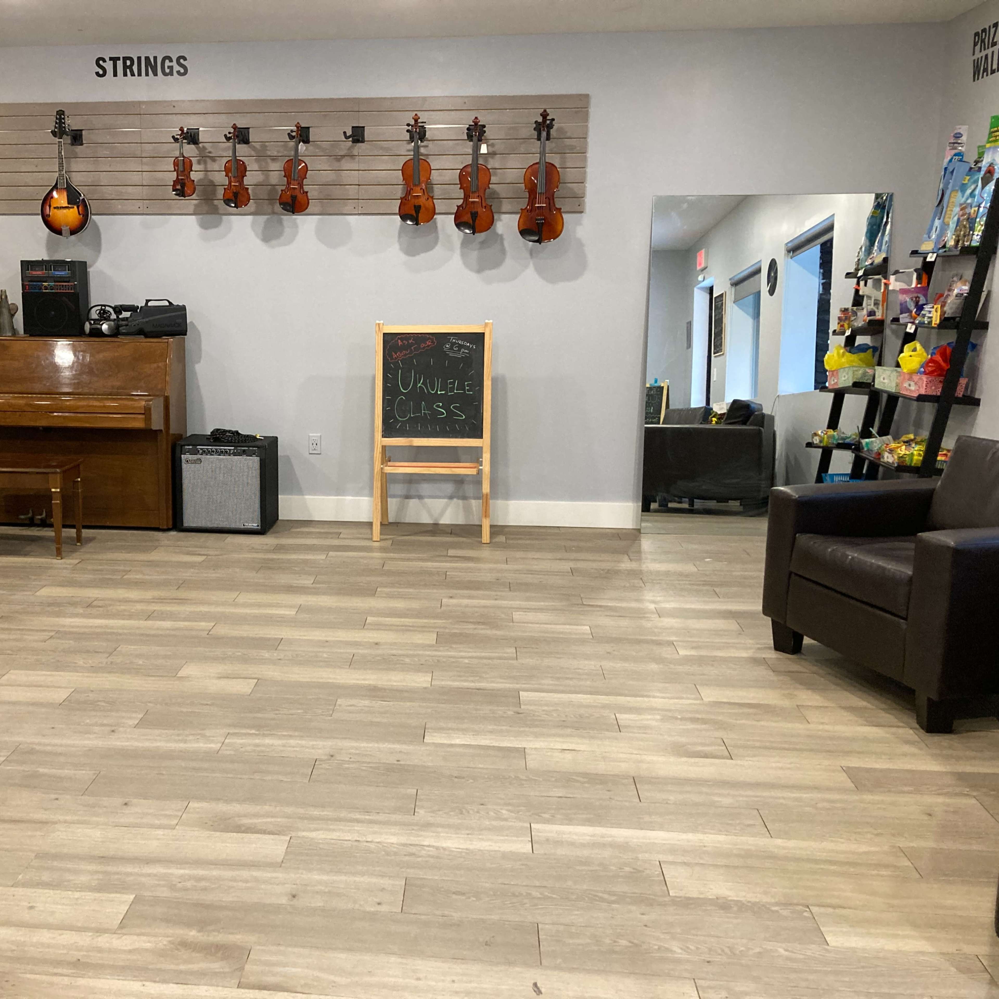 A music room with various string instruments displayed on a wall, a piano, an amplifier, and a chalkboard sign indicating ukulele classes.