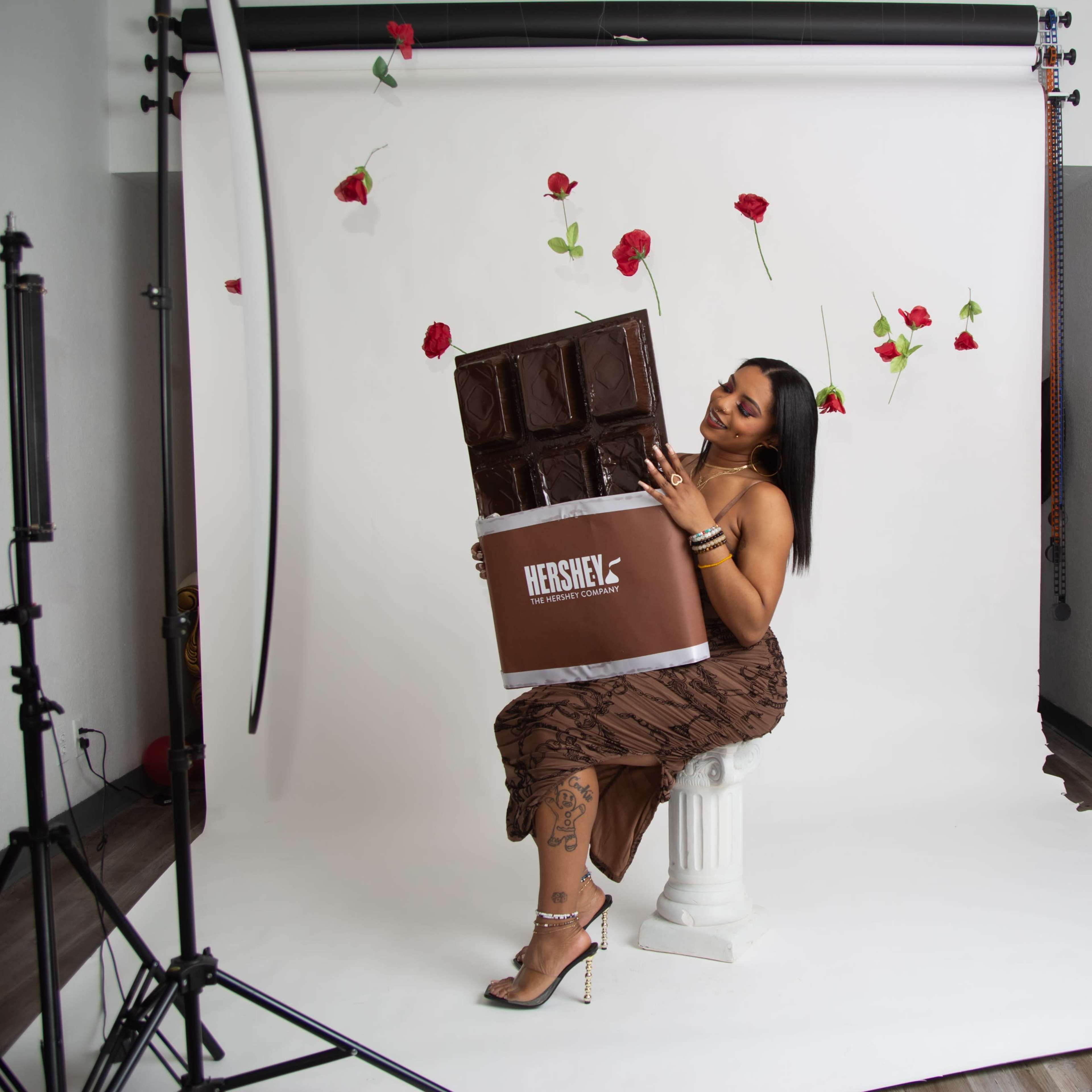 A woman holds a large Hershey's chocolate bar while sitting on a pedestal in front of a white backdrop adorned with floating roses.