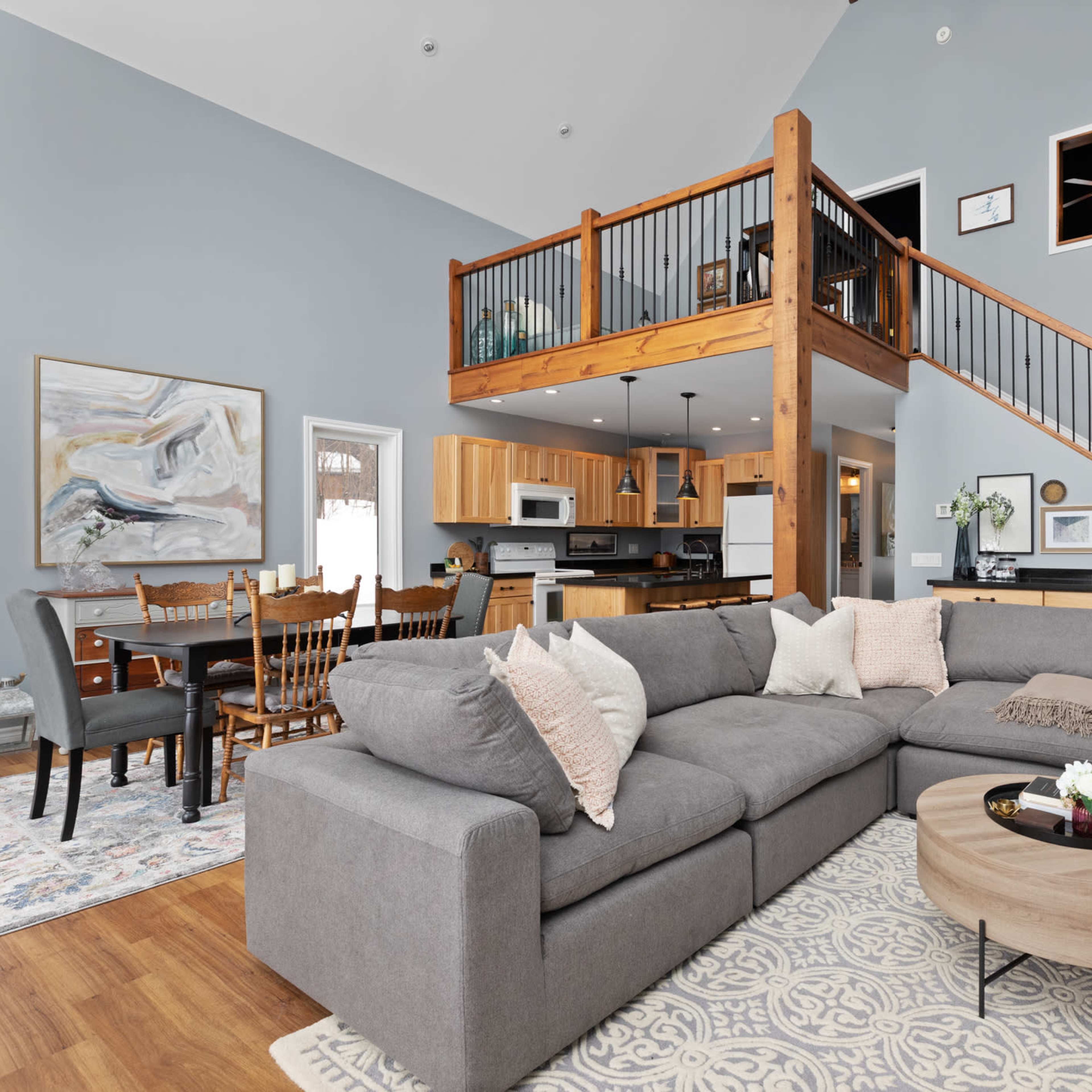 A spacious living area with a gray sectional sofa, a round coffee table, and a dining table adjacent to a staircase leading to a lofted space.