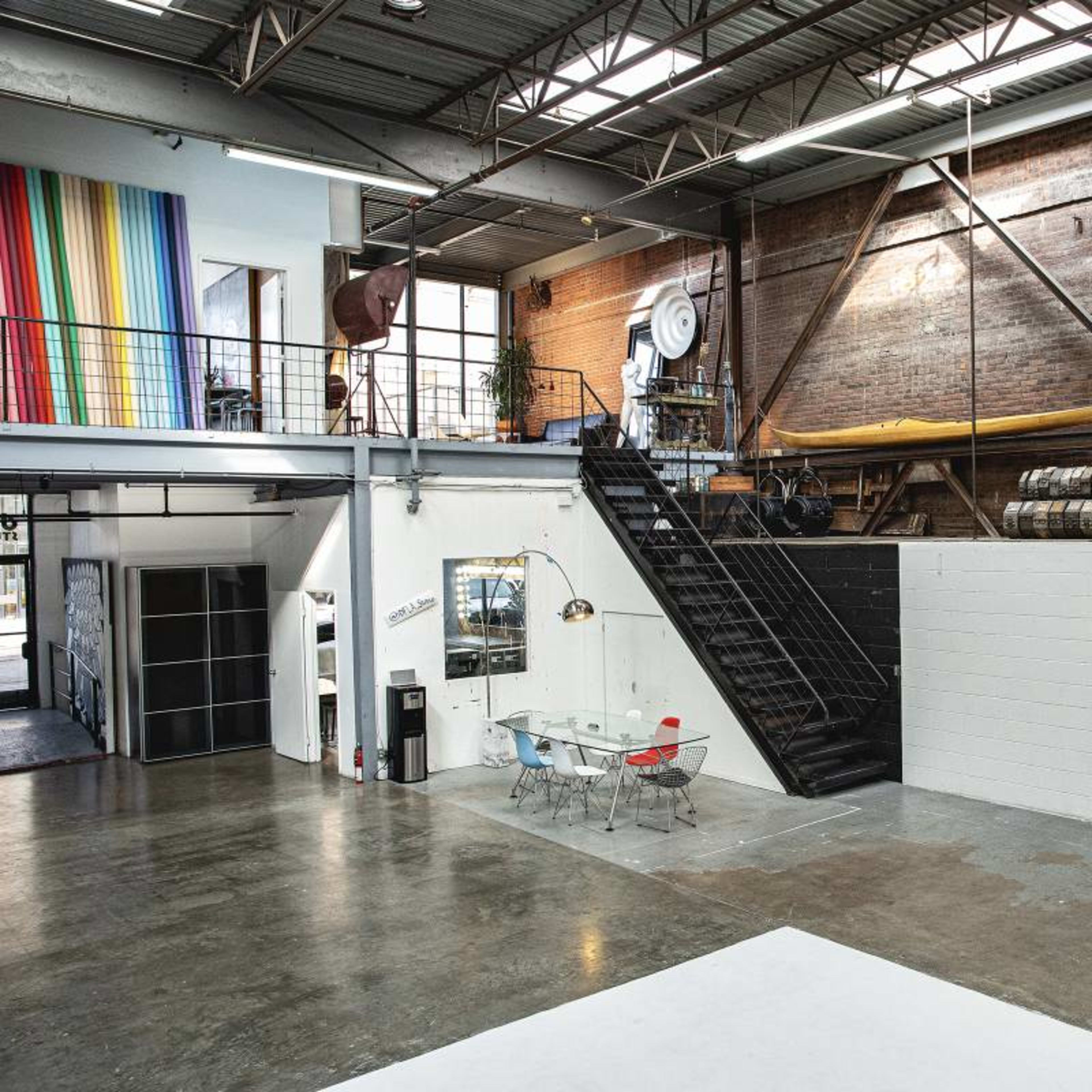 An industrial-style loft with a staircase leading to an upper level, featuring large windows, exposed brick walls, and colorful striped artwork.