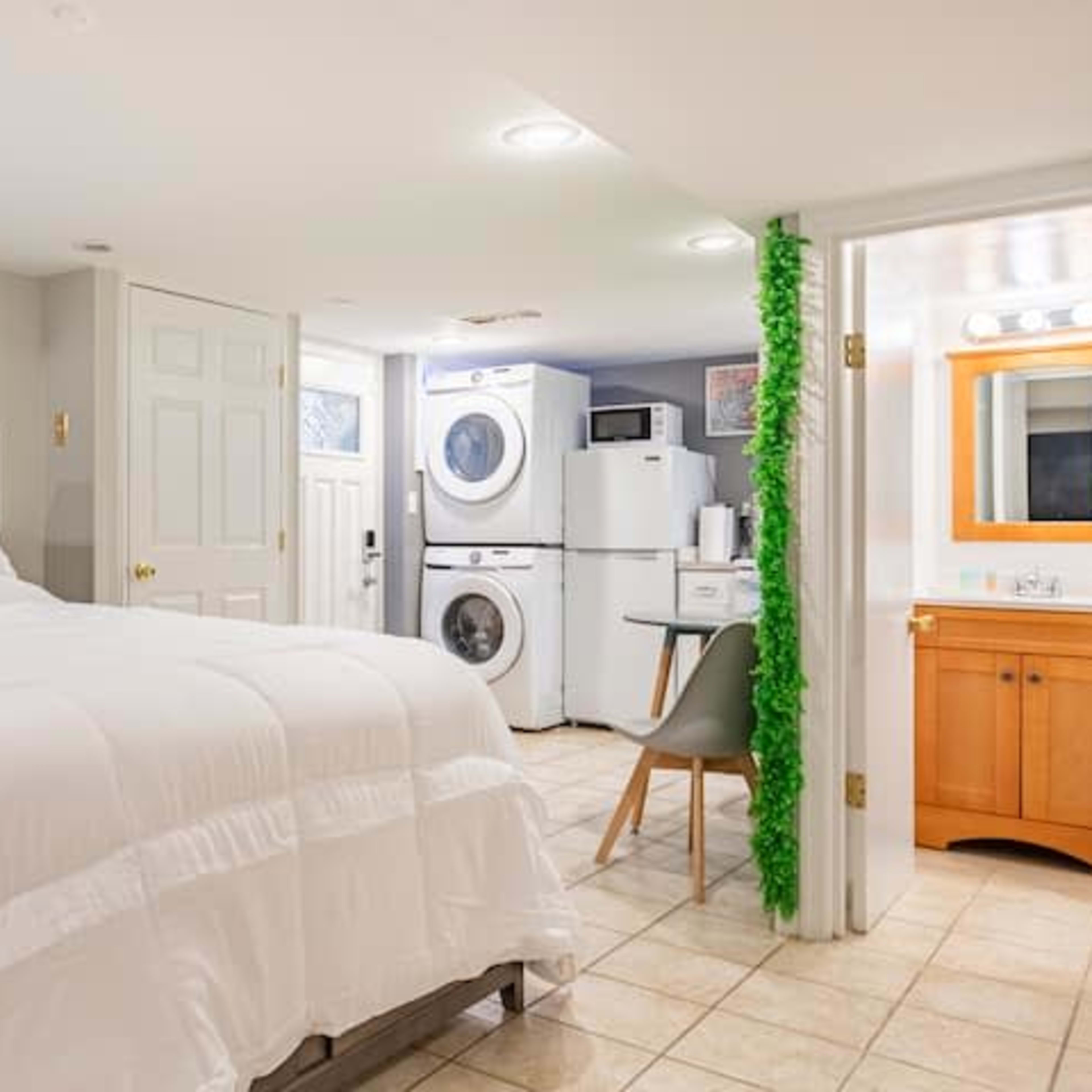 A tidy basement bedroom features a bed, a washing machine and dryer, and a bathroom area with a sink and mirror.