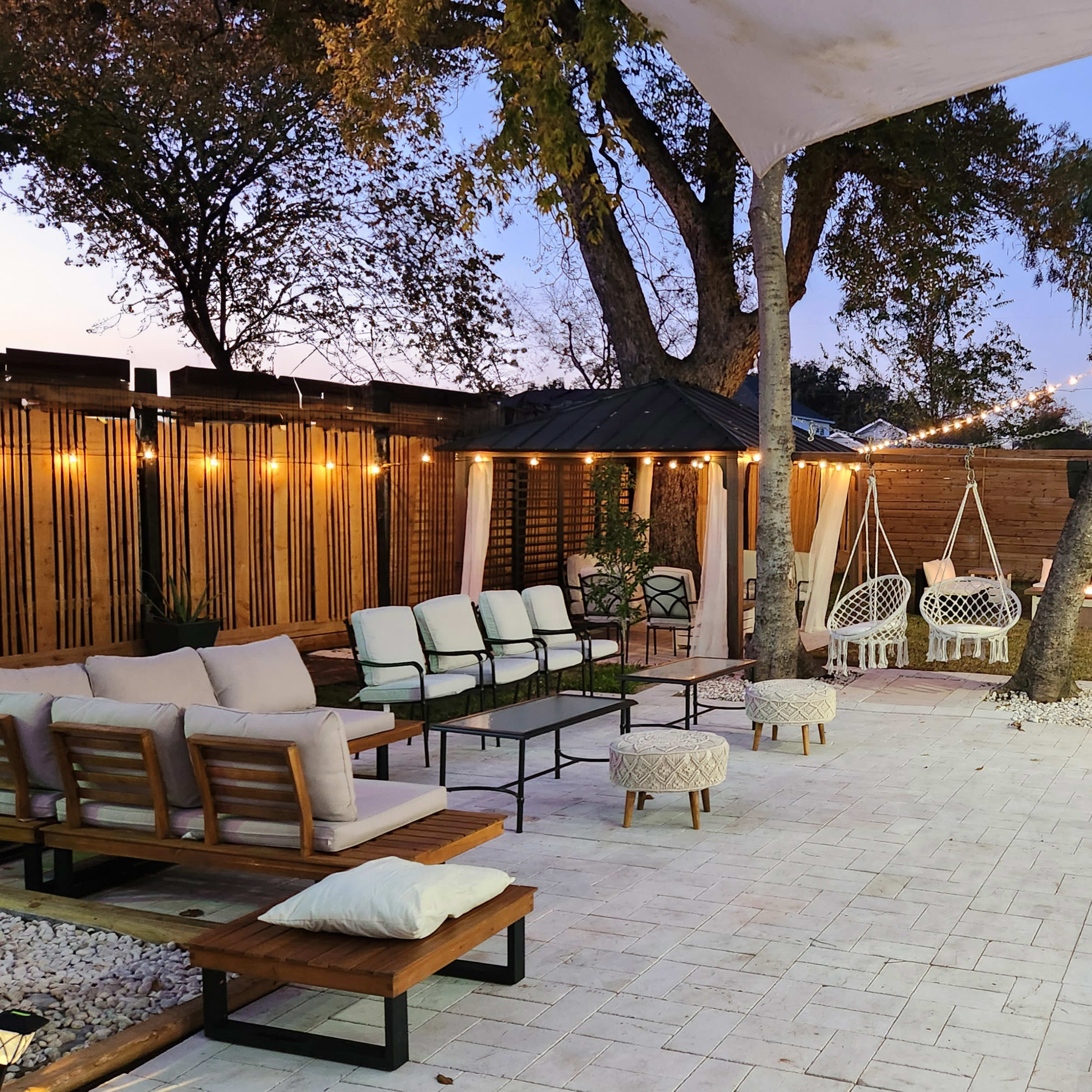 A stylish outdoor lounge area features seating arrangements, decorative lighting, and a wooden deck surrounded by trees.