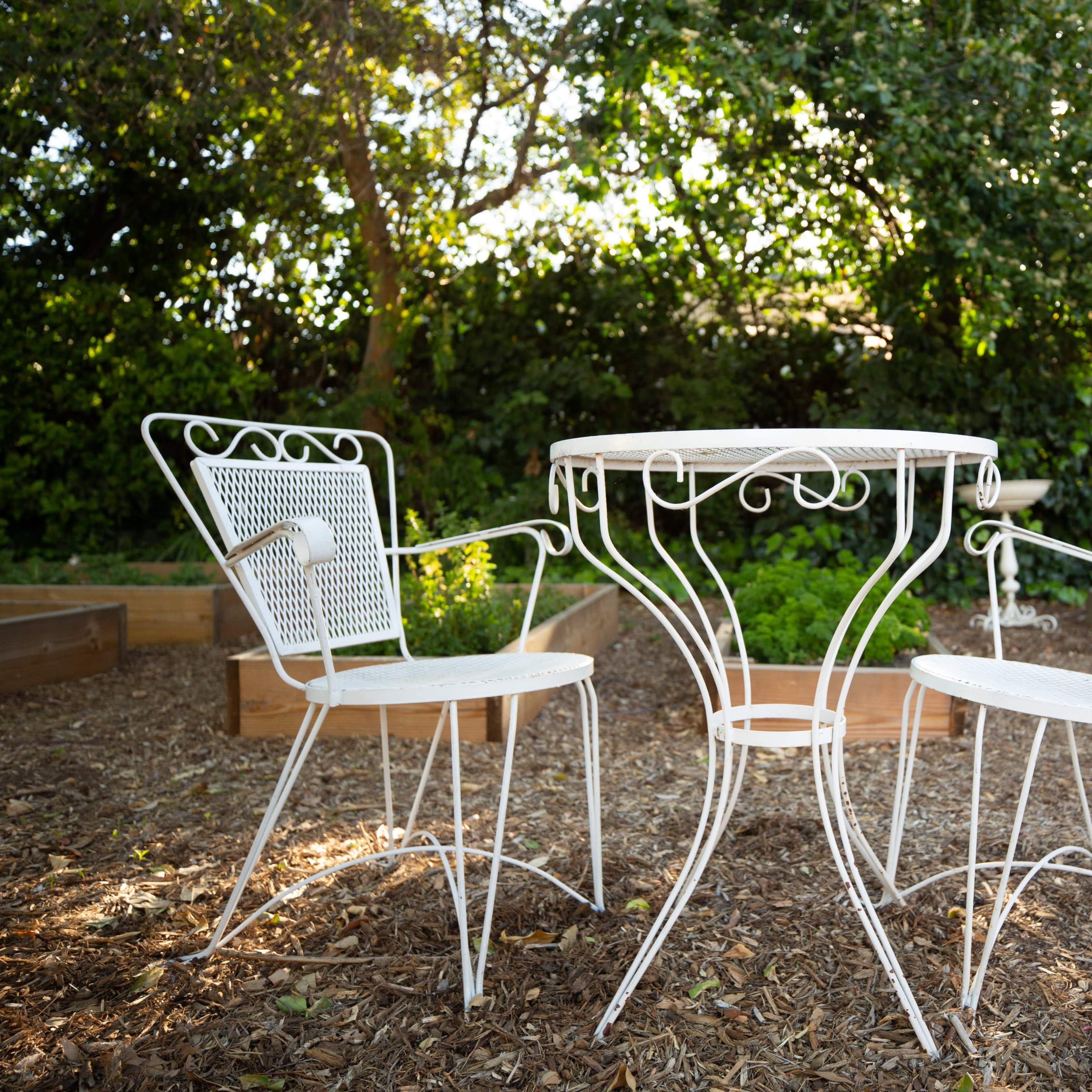 A white metal table with two matching chairs is positioned on mulch in a garden surrounded by greenery.