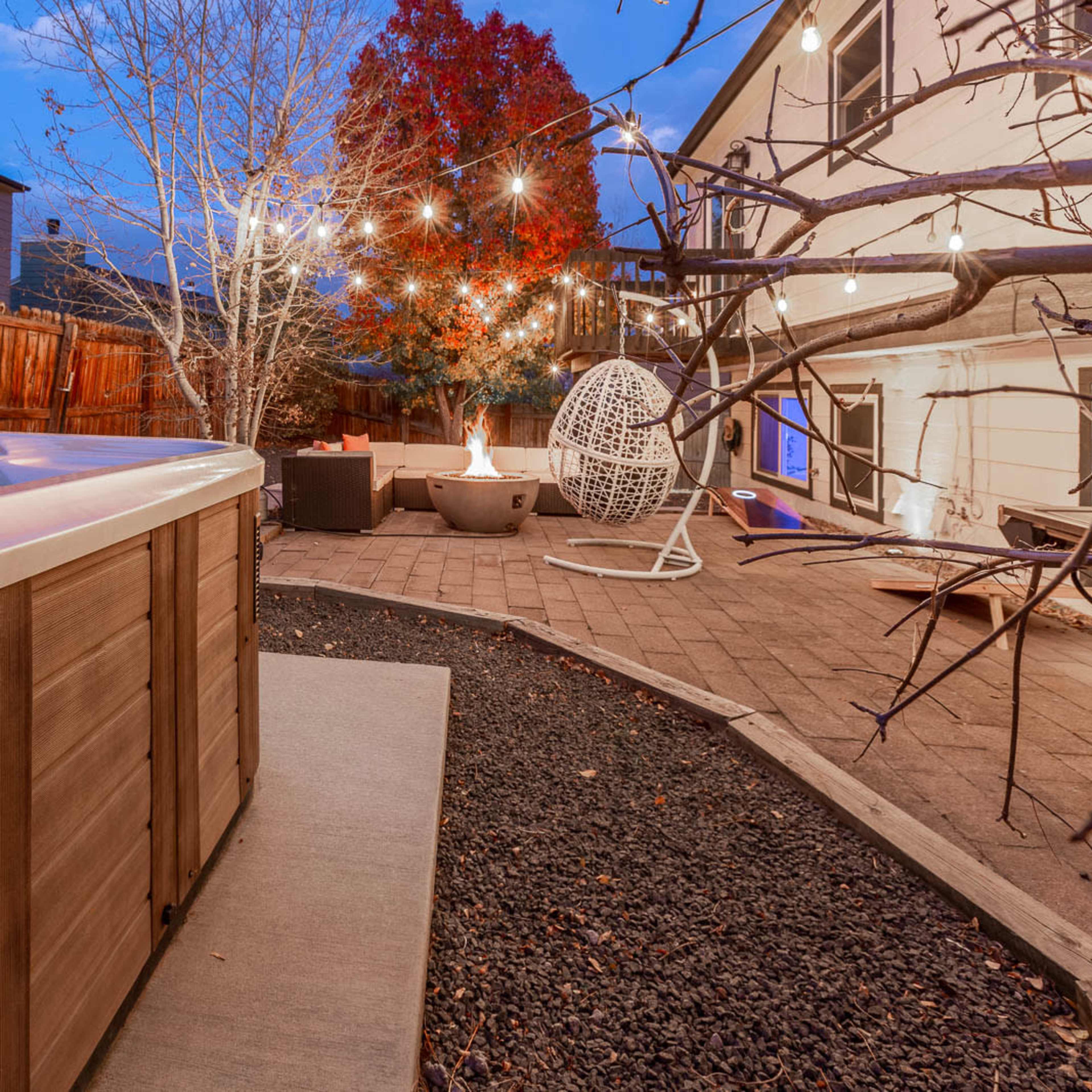 A backyard featuring a hot tub, a fire pit, and string lights, with a nearby tree displaying autumn leaves.