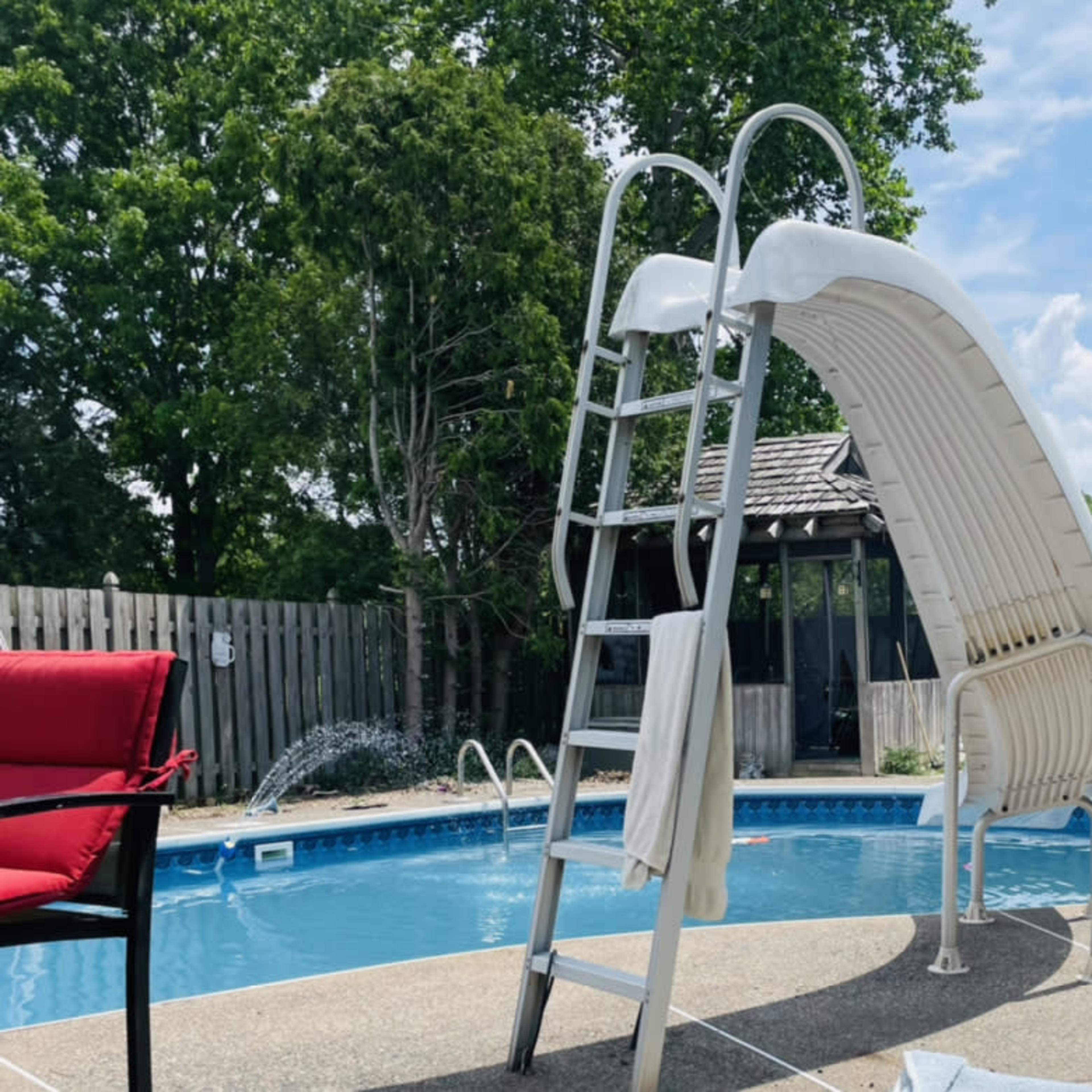 A white water slide stands beside a circular swimming pool in a backyard, with trees and seating areas in the background.