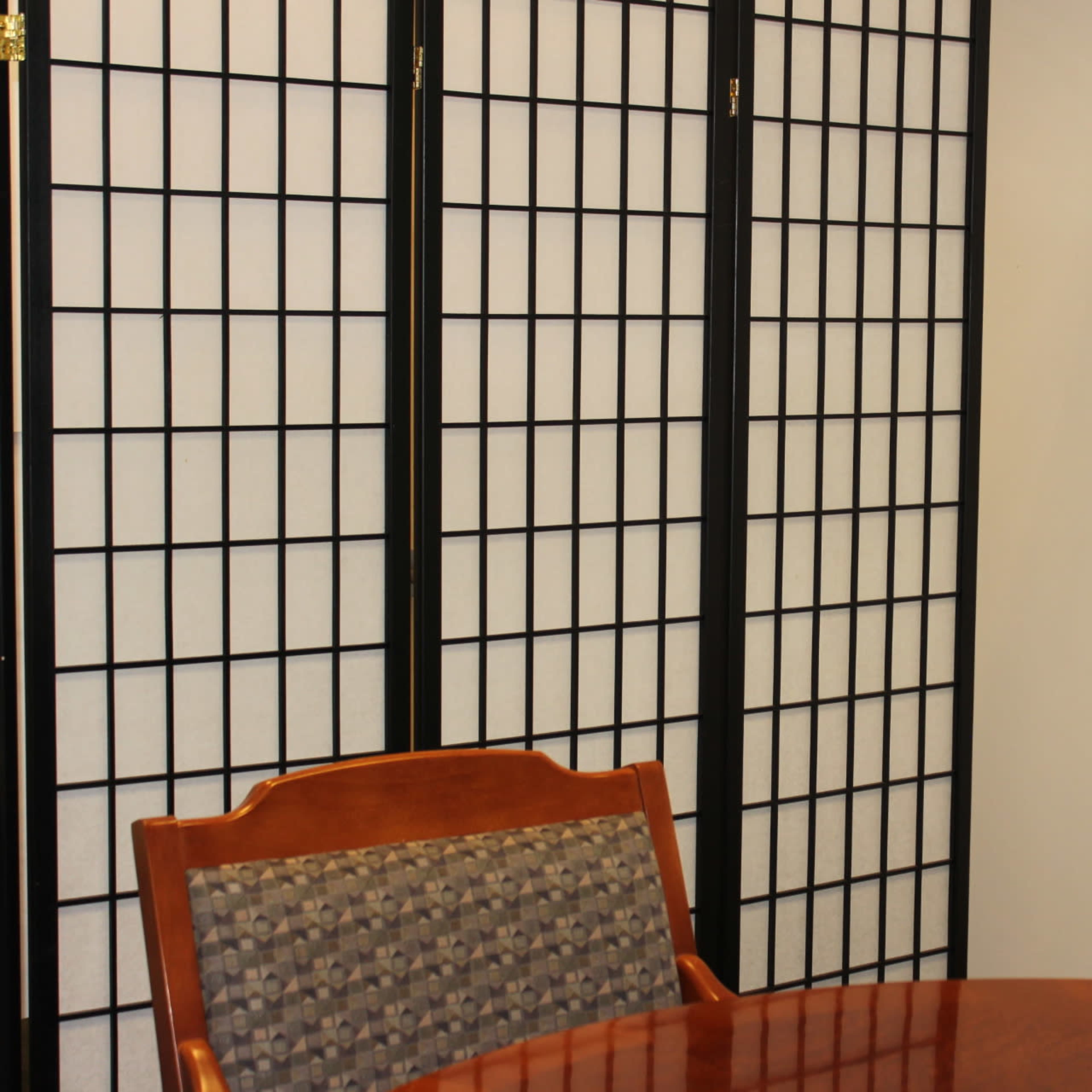 A wooden table with a patterned chair is set near a folding screen in a room with a plain white wall.