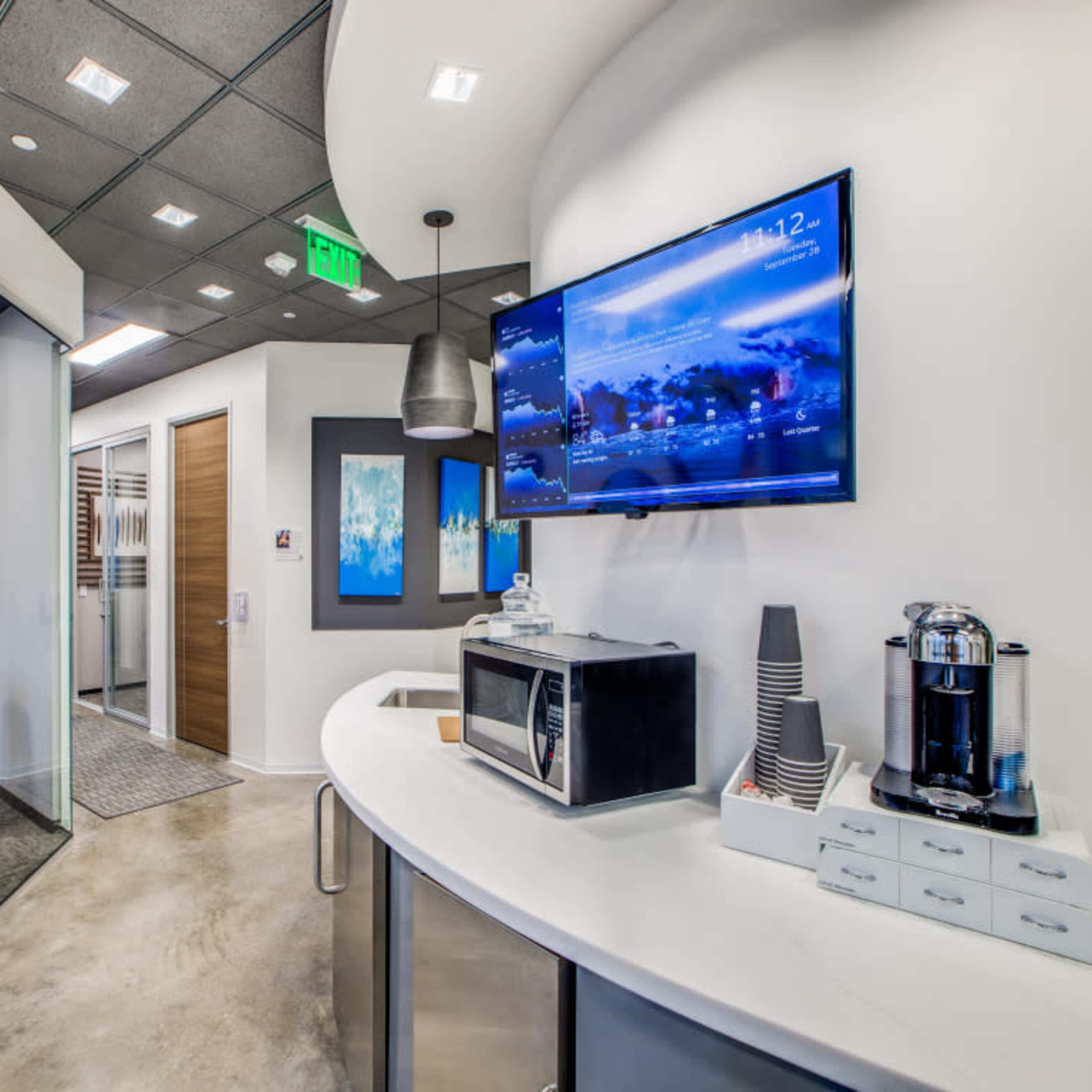 A modern office kitchenette area with a microwave, coffee maker, and a large monitor displaying a digital screen.