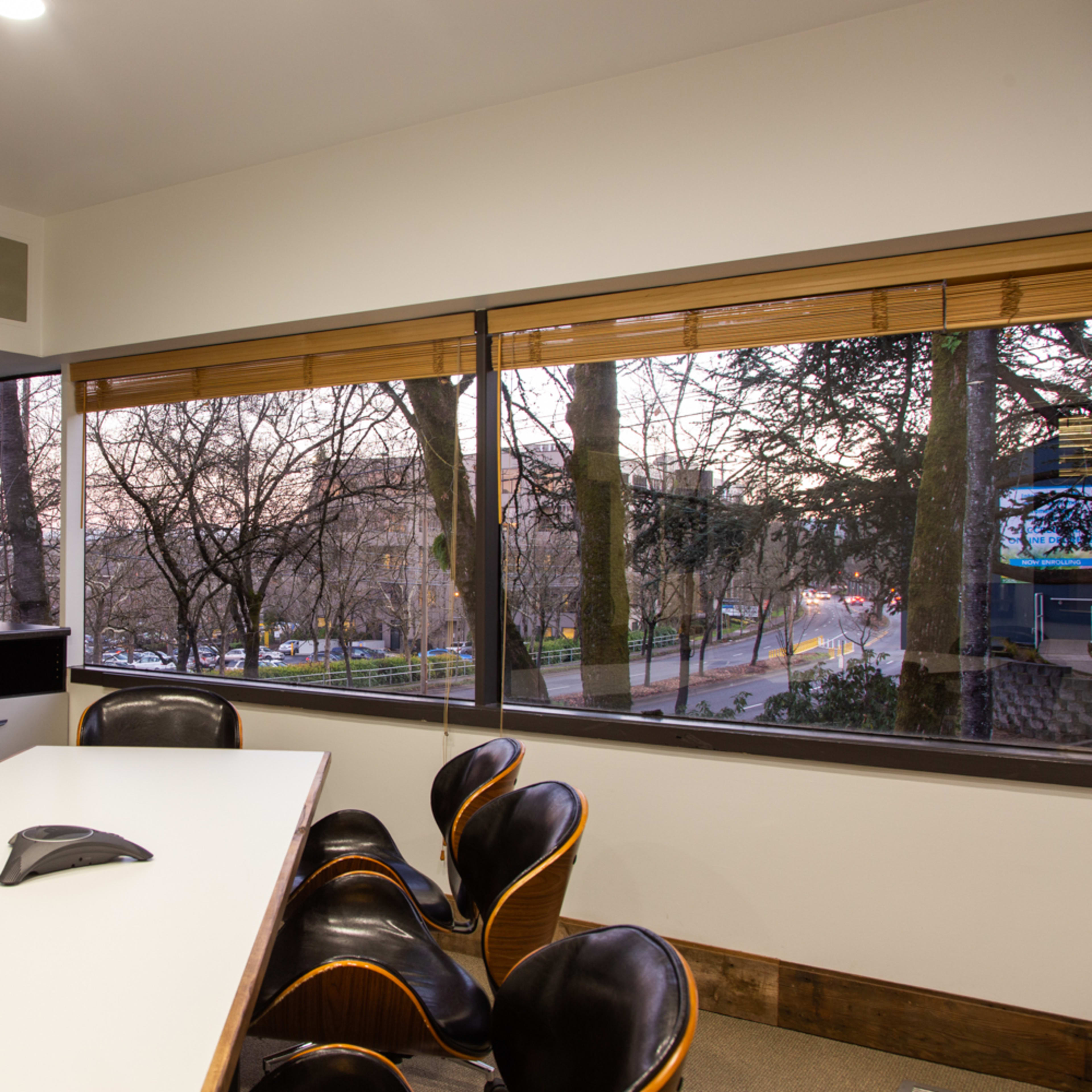 A conference room features a long table with black chairs and a large window overlooking a tree-lined street.